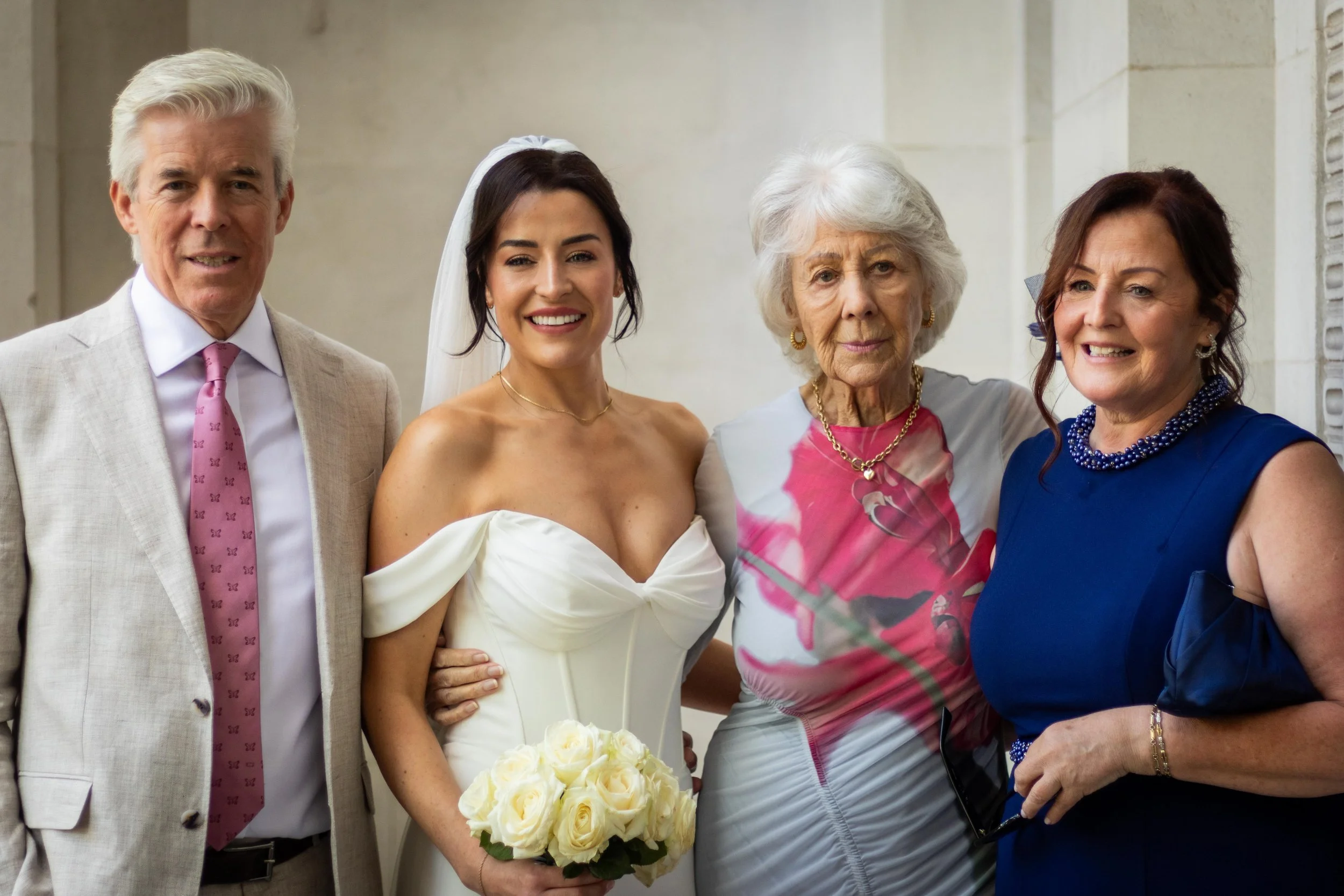 Group of five people at a wedding, including a bride in a white dress holding a bouquet of white roses, standing with three women and one man, all dressed formally.