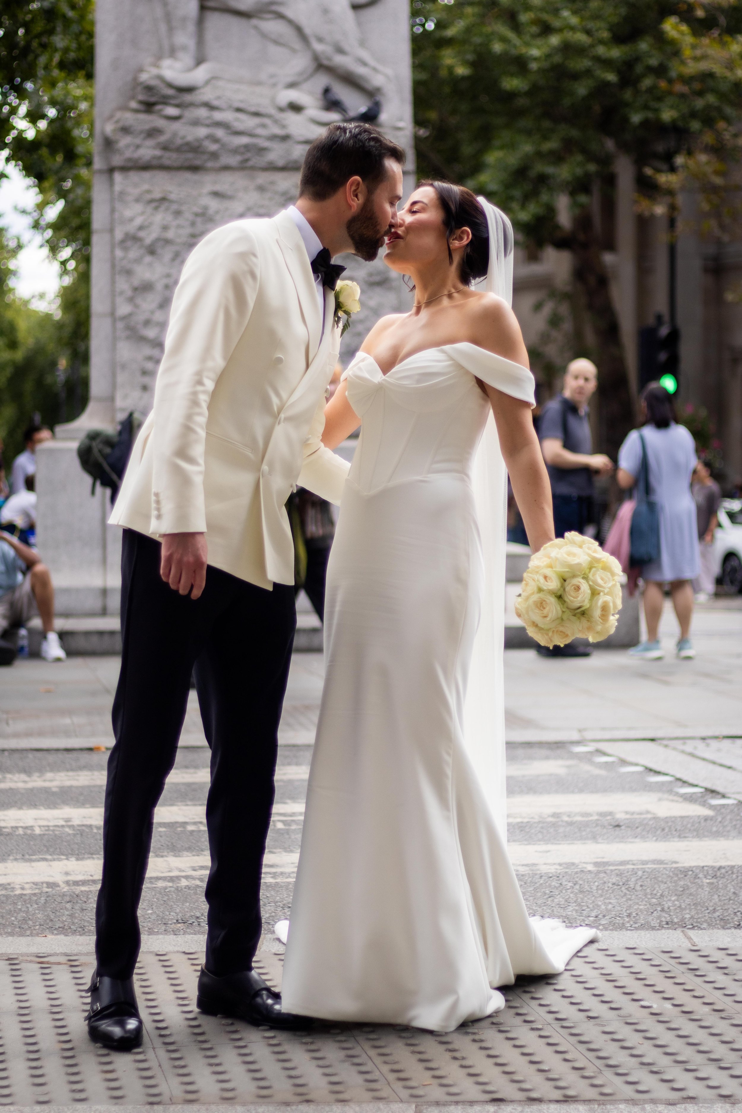 A bride and groom in wedding attire sharing a kiss on a city street. The bride is holding a bouquet of white roses in her left hand, and they are standing near a statue and some pedestrians are in the background.