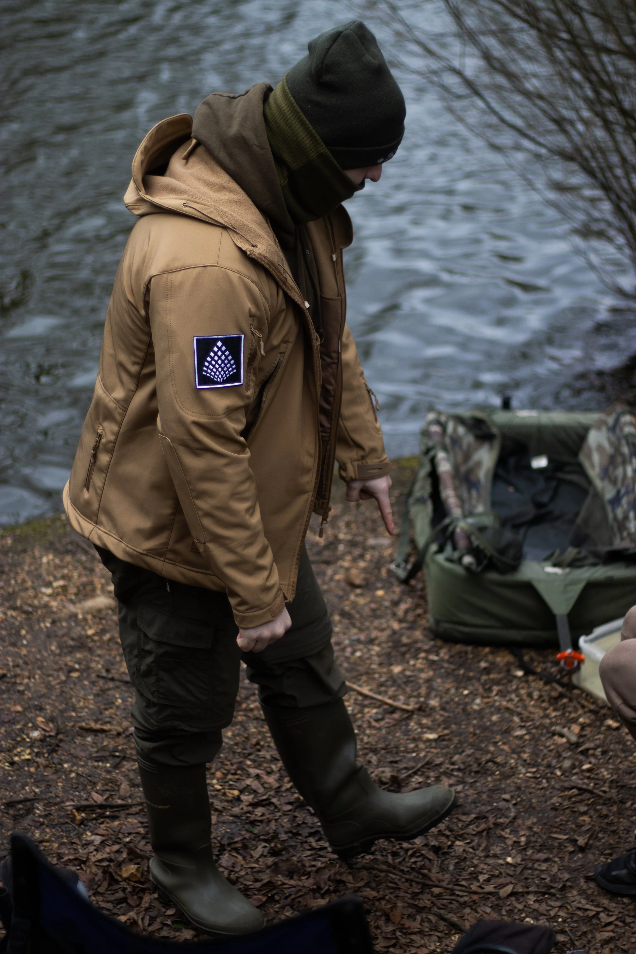Person dressed in outdoor gear, including a hooded jacket, beanie, and rubber boots, standing by a body of water with gear on the ground nearby.