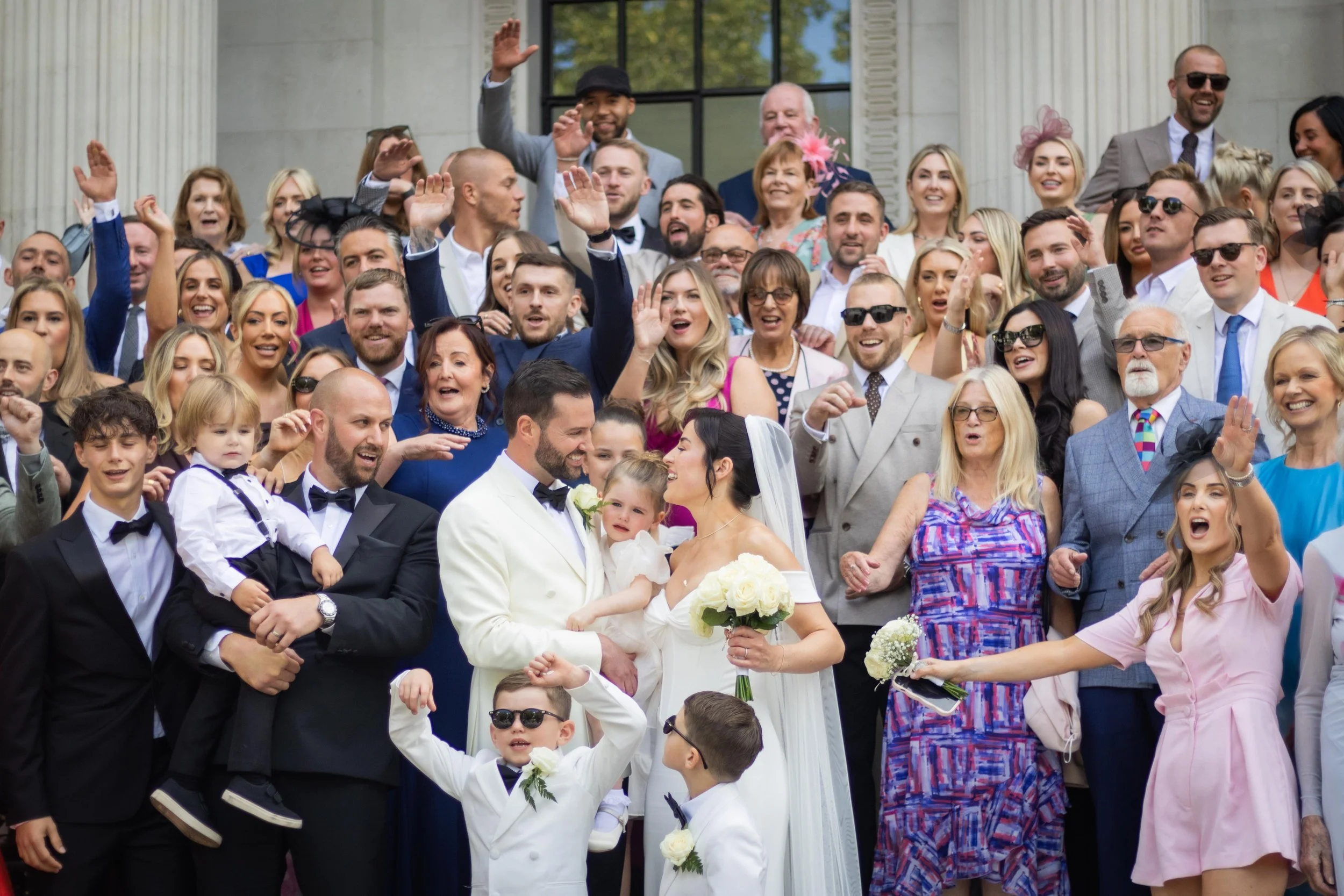 A large group of people gathers on the stairs of a building for a wedding celebration, with a bride and groom at the center surrounded by family and friends.