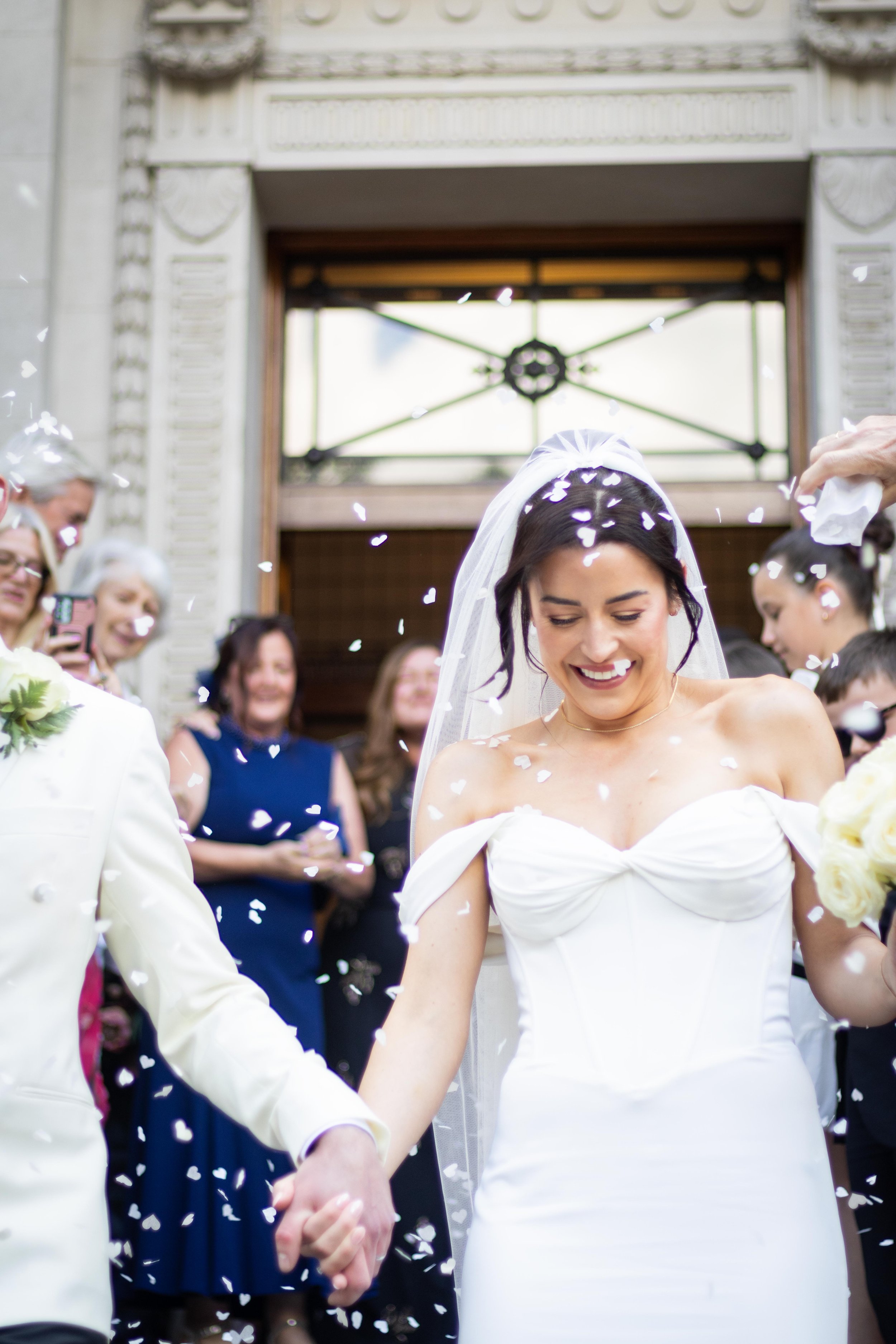 Bride and groom holding hands after wedding ceremony, surrounded by guests, with confetti falling in front of a building with ornate architectural details.