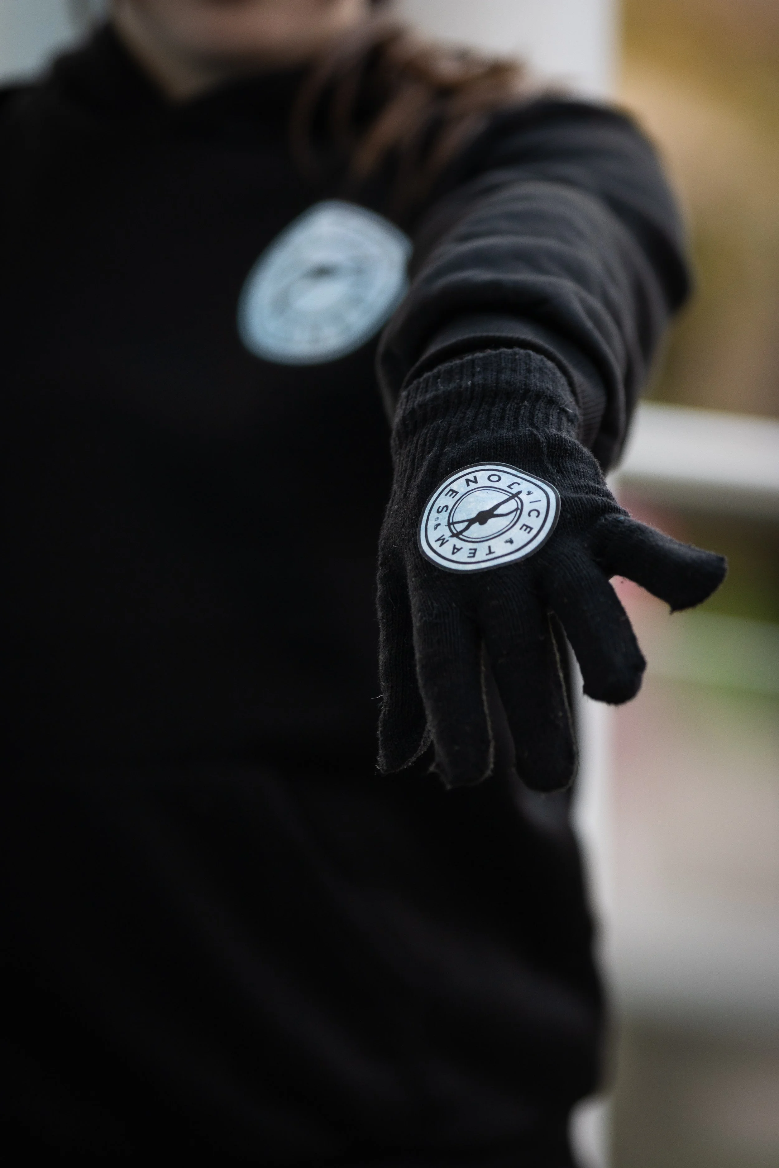 Close-up of a person wearing black gloves and a black long-sleeve shirt, with a circular logo on the glove and shirt that reads 'TECNOLOGISM' and features a compass needle.