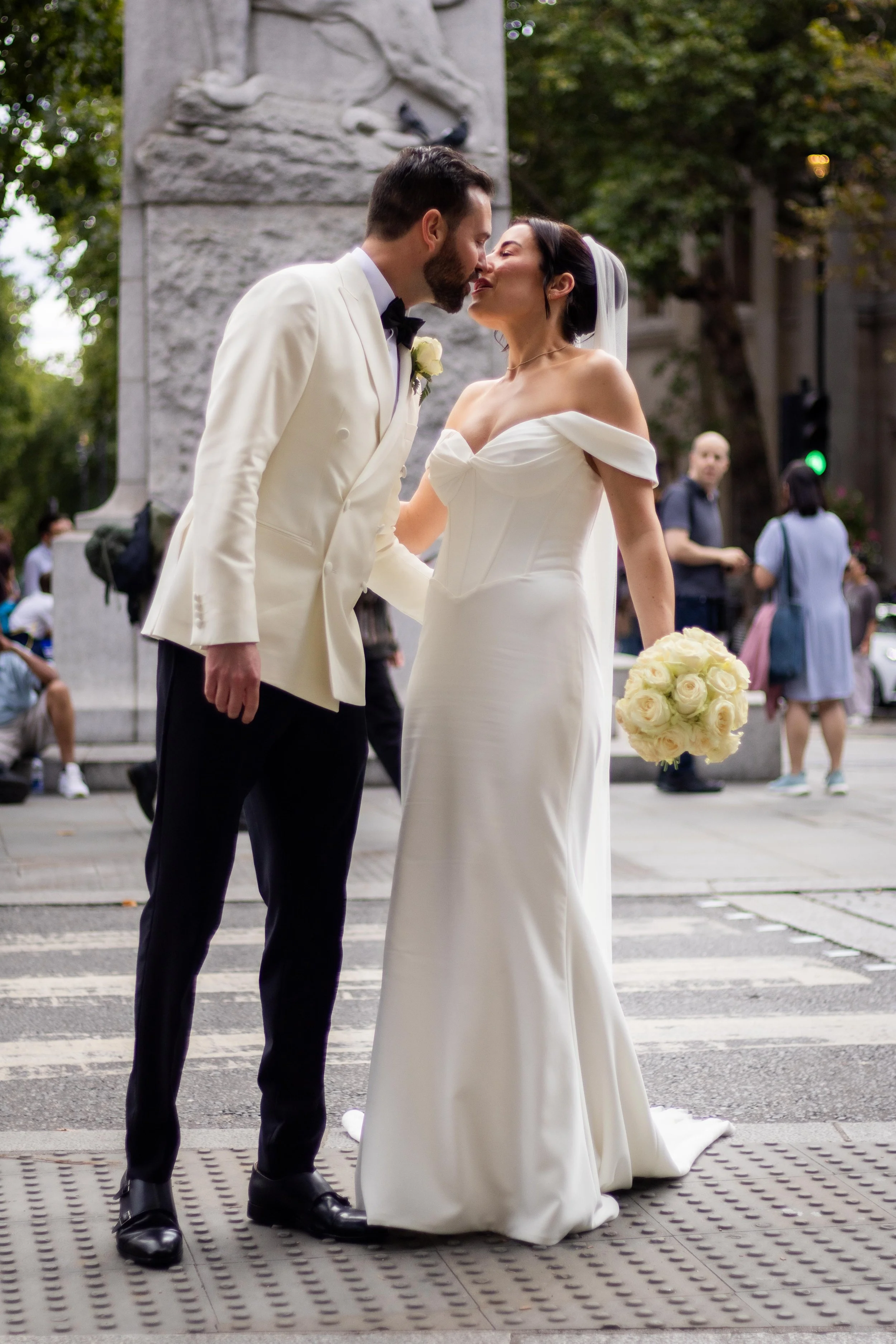 A bride and groom sharing a kiss on their wedding day in an urban setting, with the bride holding a bouquet of white roses and wearing a white off-the-shoulder gown, and the groom in a white tuxedo jacket and black pants, surrounded by onlookers.
