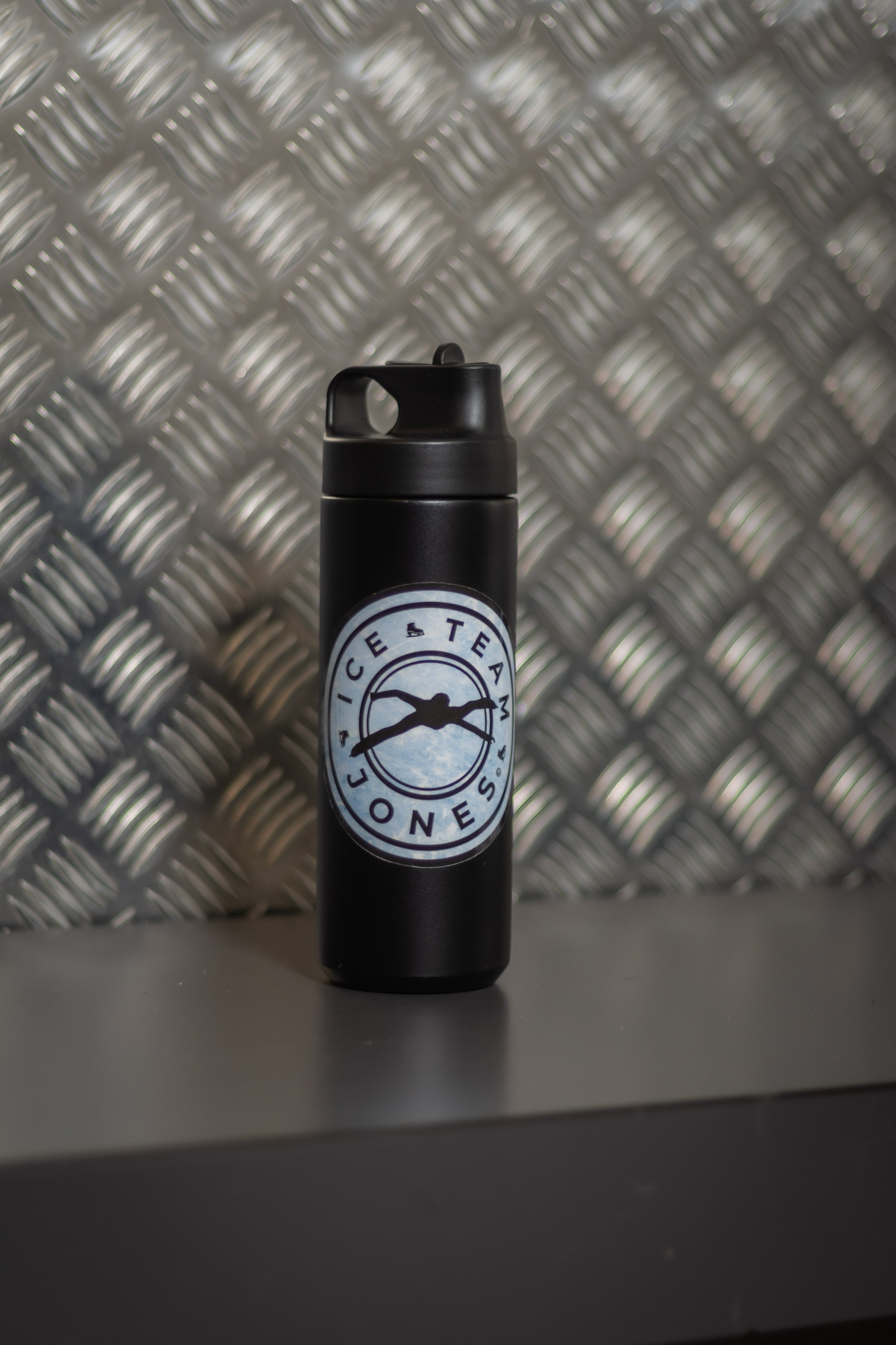 A black water bottle with a logo that says 'Ice & Tea Jones' and features a mountain silhouette, placed on a gray surface with a metal patterned background.