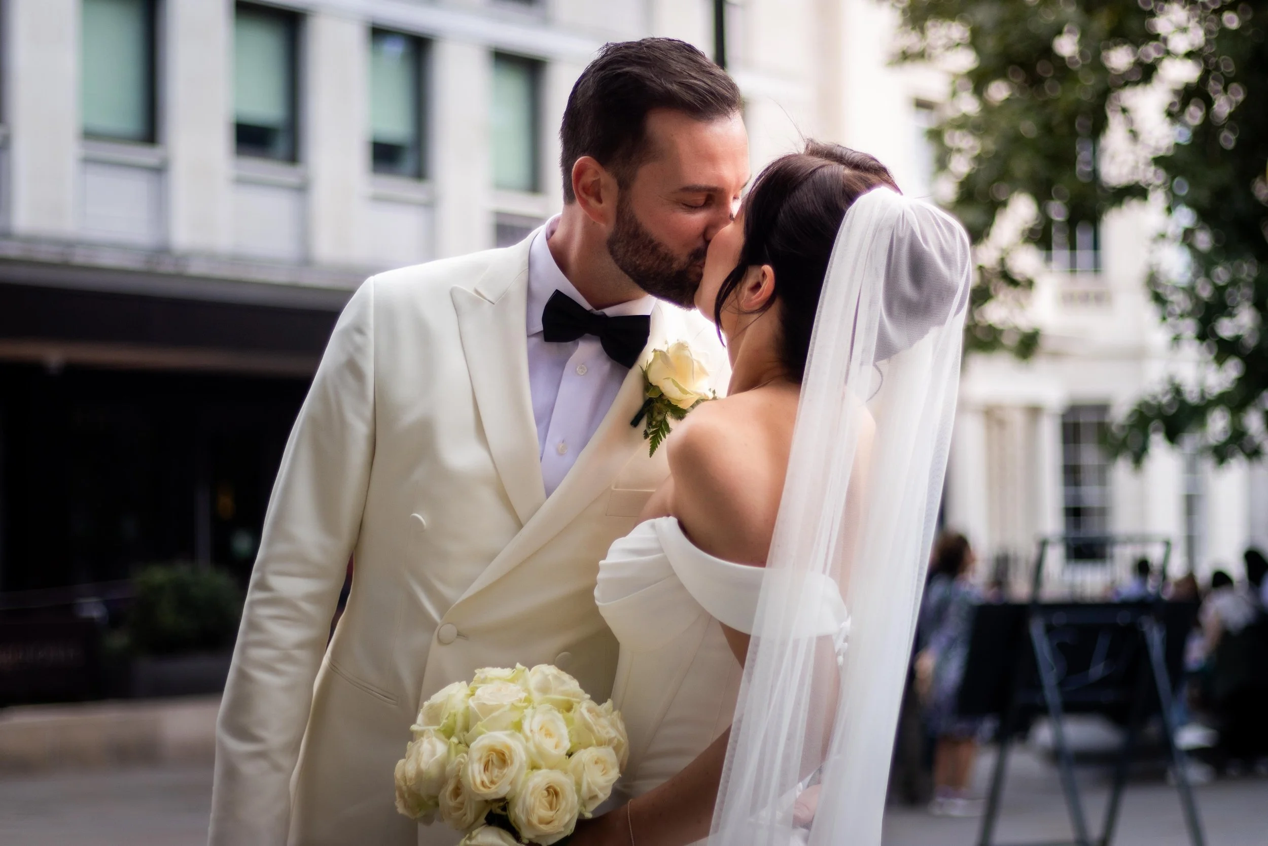 A newlywed couple sharing a kiss outdoors, the groom dressed in a white tuxedo with a black bow tie and holding a bouquet of white roses, and the bride in a white off-the-shoulder wedding gown with a veil.