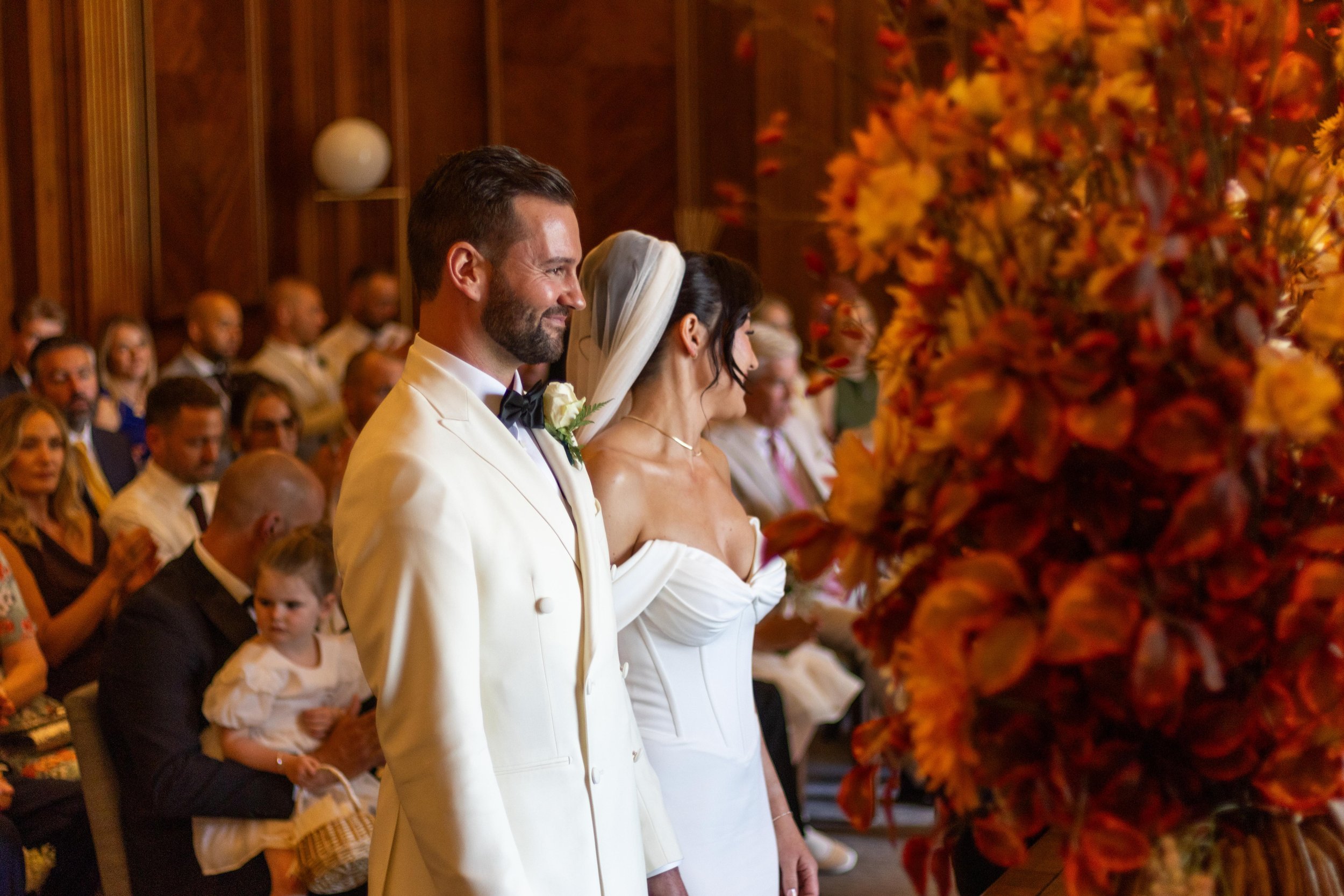 A bride and groom standing during a wedding ceremony, surrounded by seated guests, with large floral arrangements in the foreground.