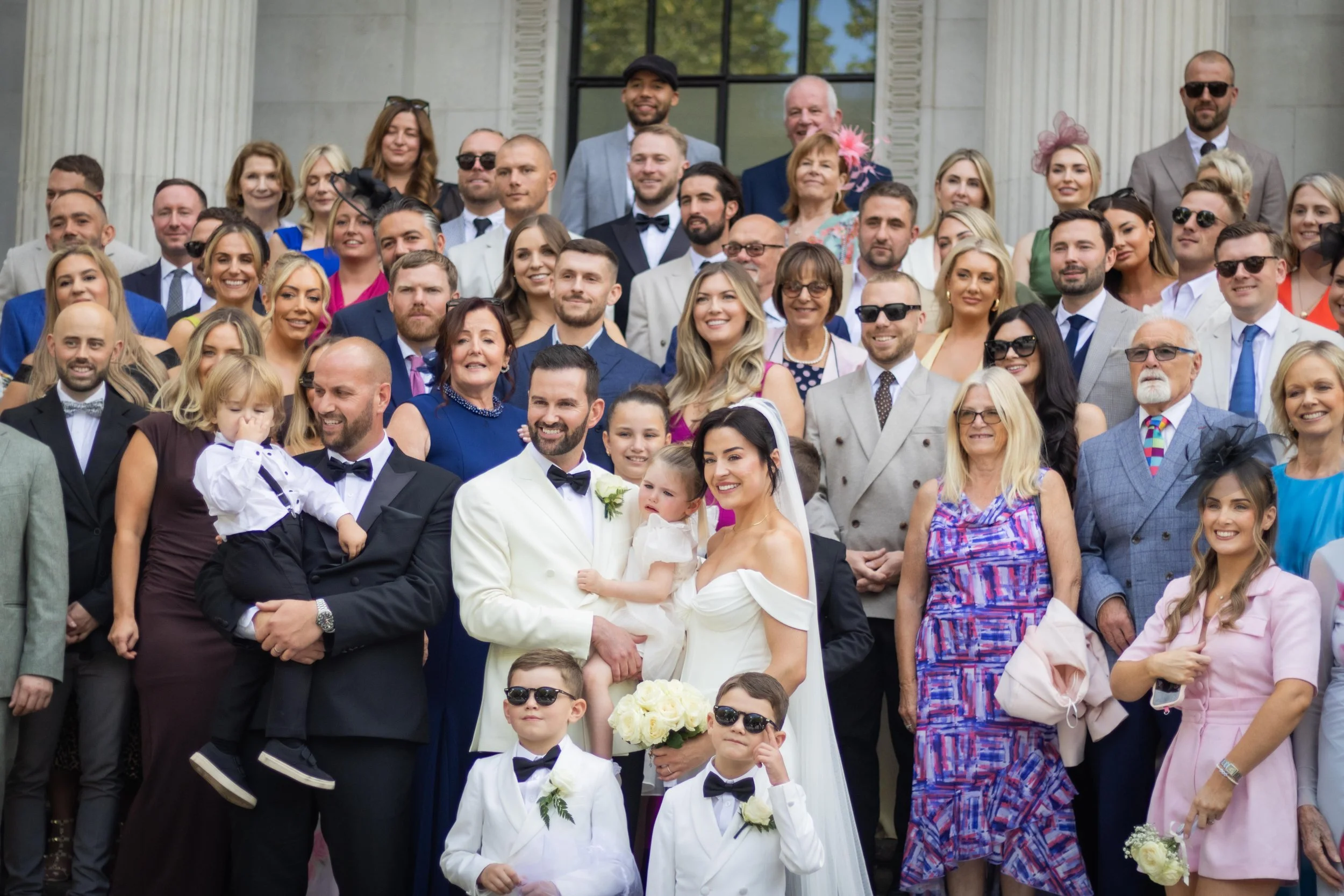 Group photo of wedding guests and family on the steps of a building, including bride and groom, children, and adults dressed in formal attire.