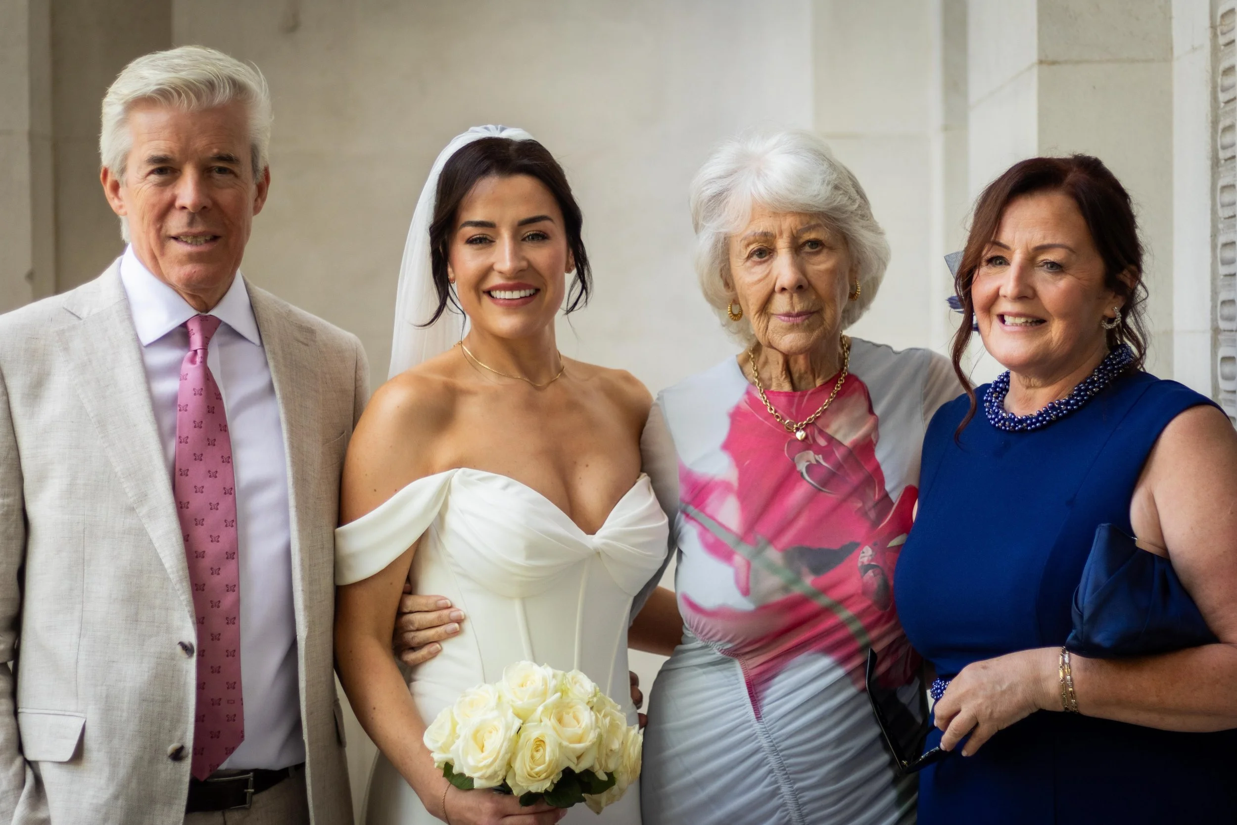 Group of five people, including a bride in a white wedding dress holding a bouquet of white roses, standing under a building with light-colored stone walls.