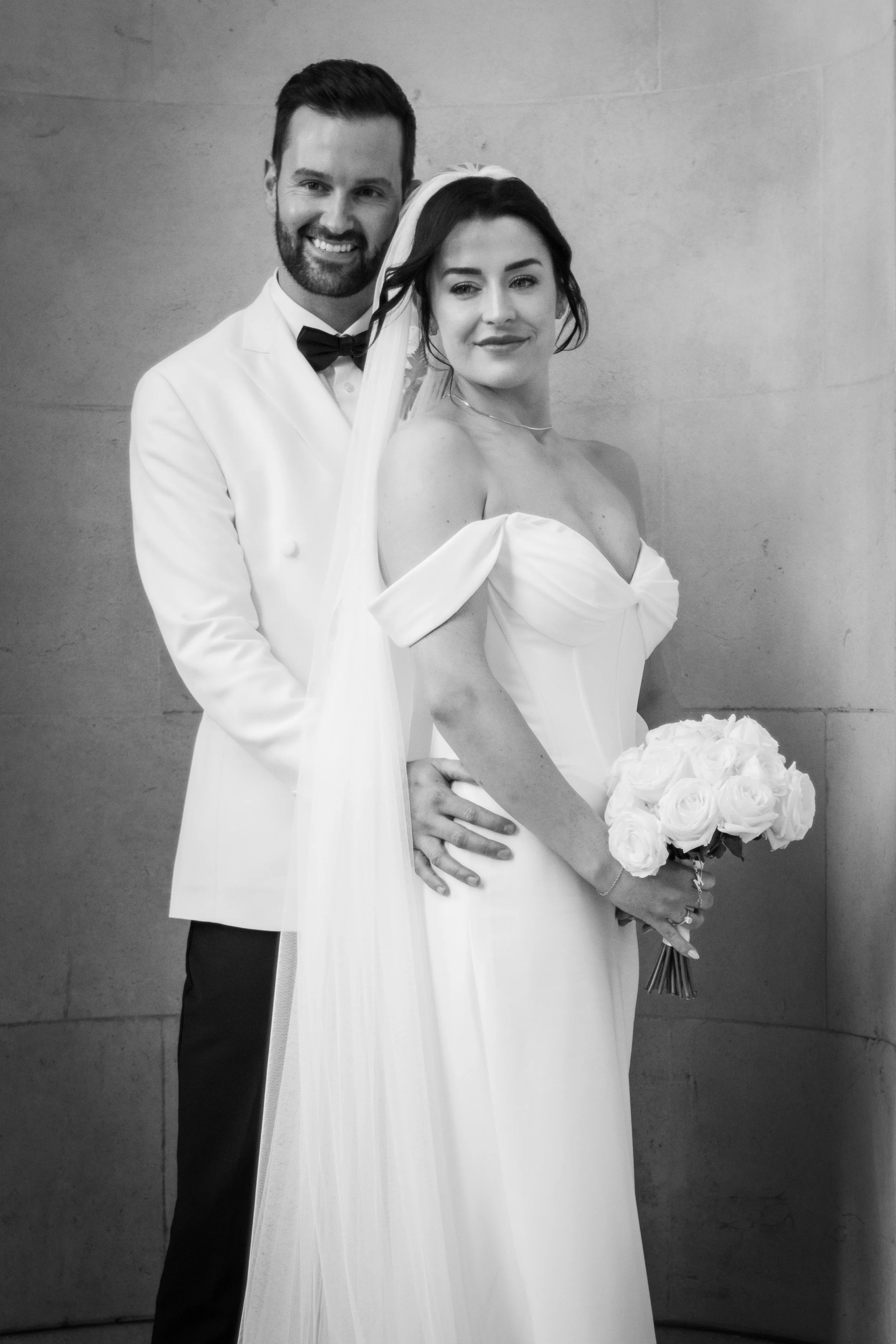 Black and white photo of a bride and groom on their wedding day. The bride is holding a bouquet of roses, and the groom is standing behind her with his hand on her waist. They are dressed in formal wedding attire, smiling softly.