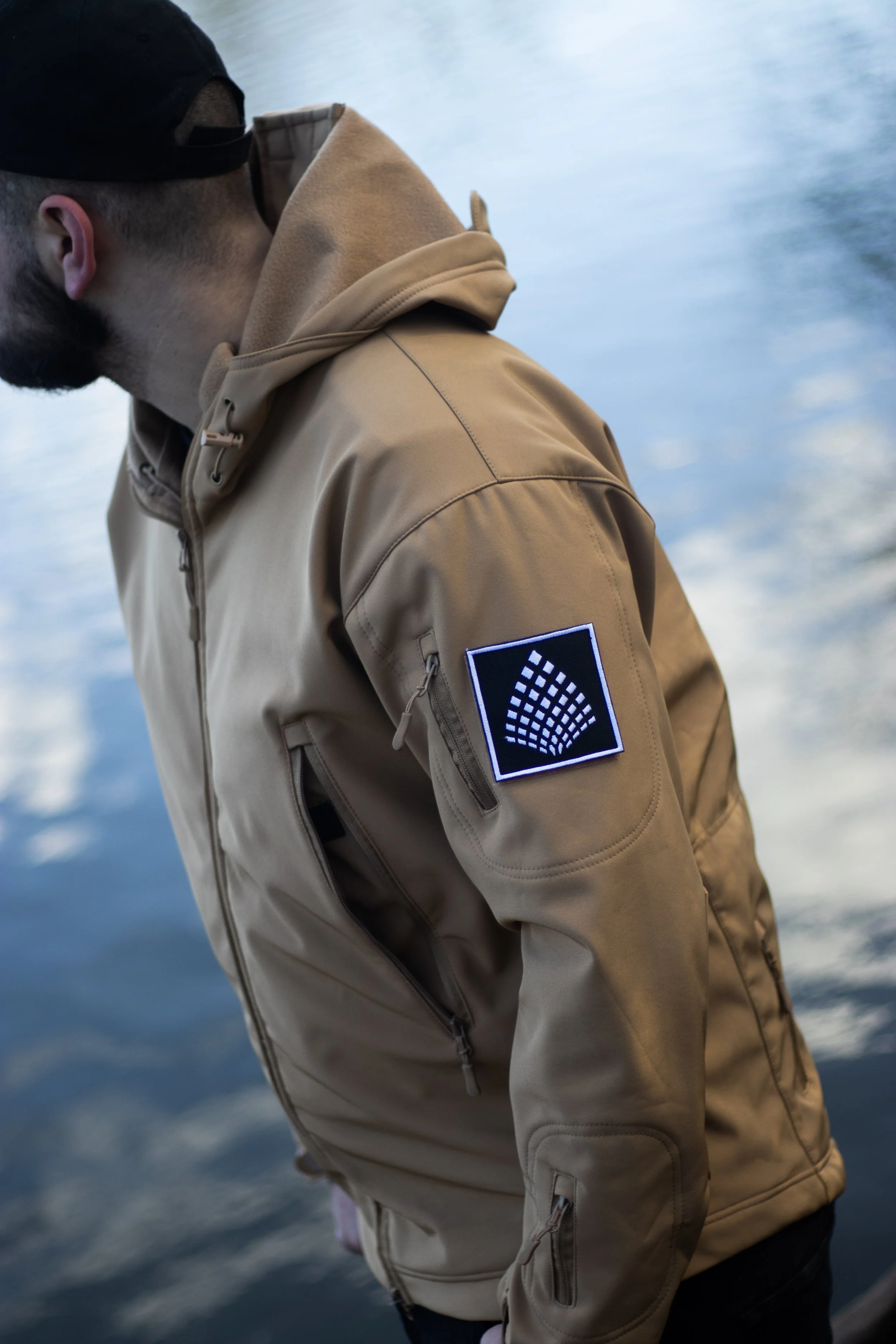 Man in beige jacket with black and white patch on sleeve, standing by water.