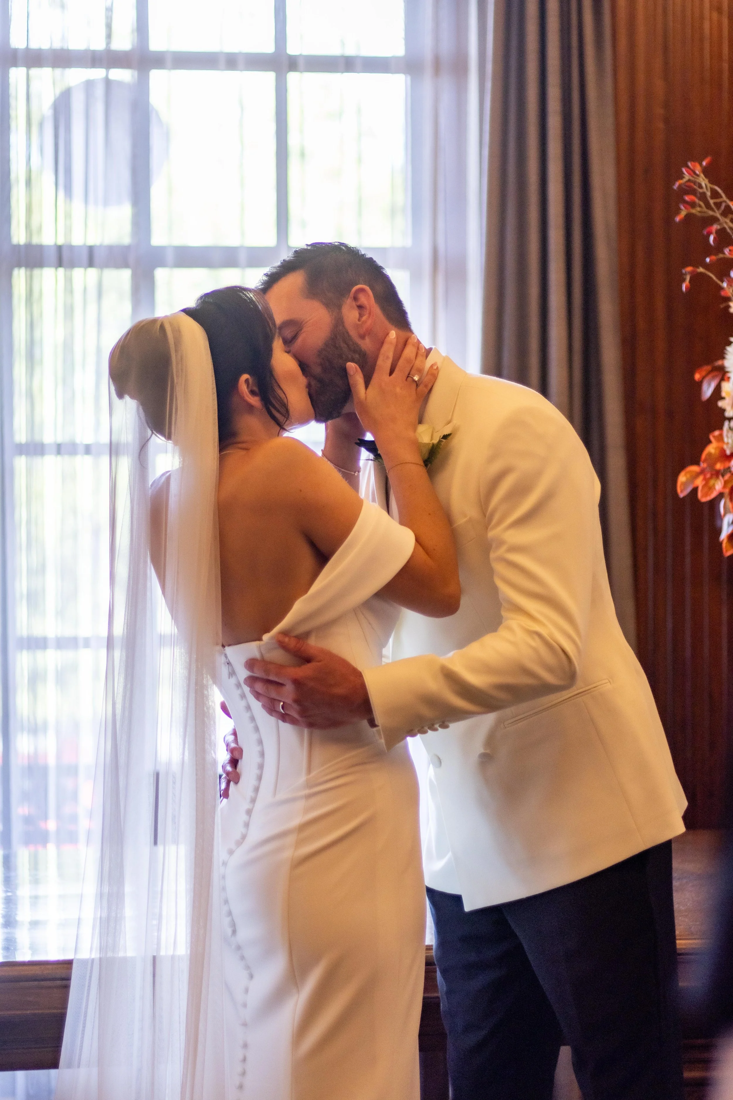A bride and groom share a kiss during their wedding ceremony, with natural light streaming through large window behind them.