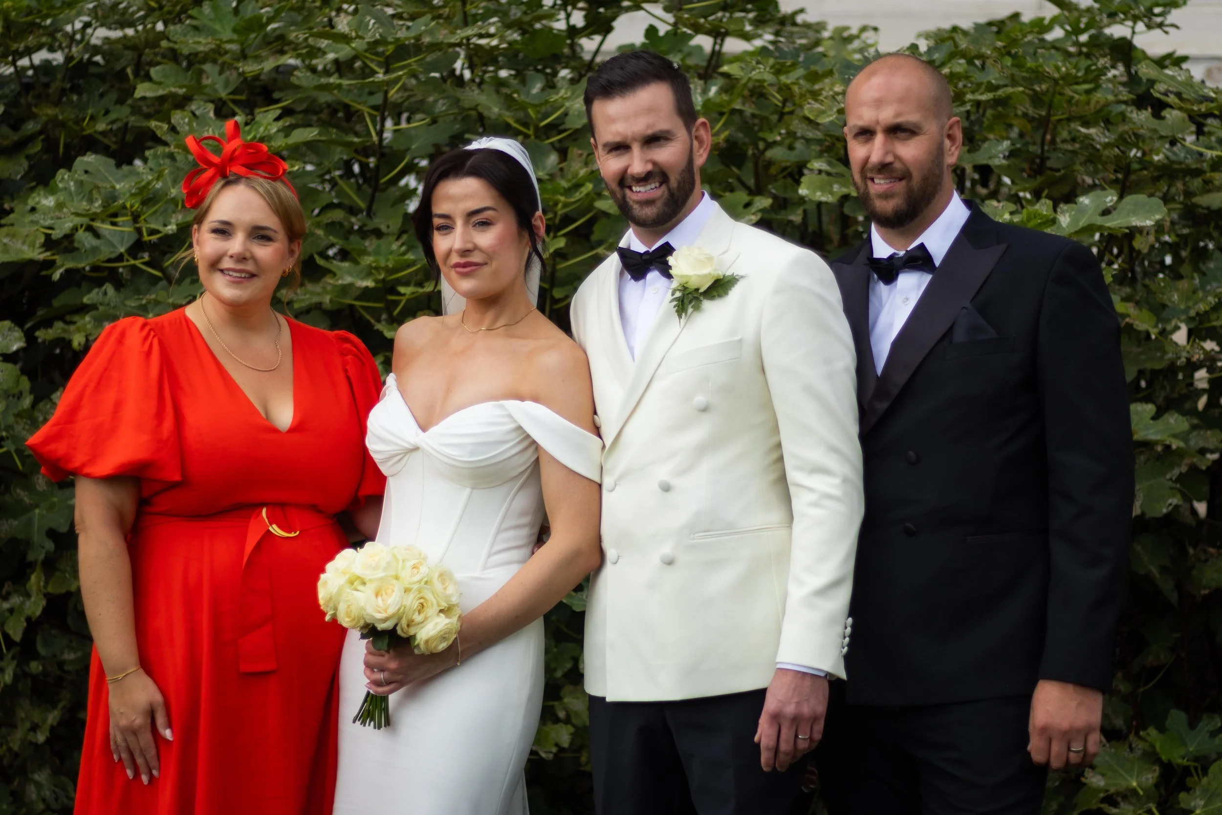 Group photo of five people dressed in formal wedding attire, standing outdoors in front of greenery. The bride is in a white strapless dress holding a bouquet of white roses. The groom beside her is wearing a white tuxedo jacket with a black bow tie.