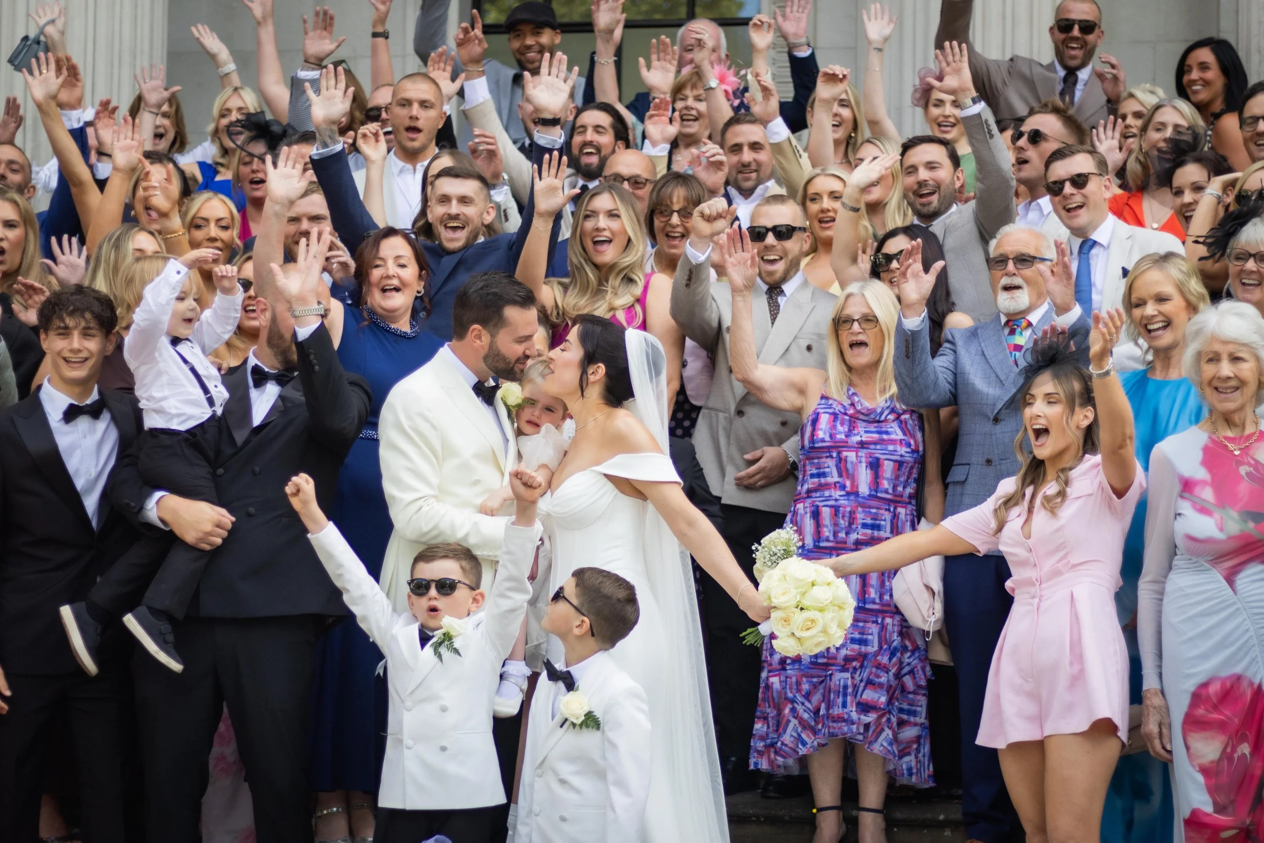 A wedding celebration with a large group of guests smiling, cheering, and raising their hands, with a bride and groom in the center sharing a kiss. The bride is holding a bouquet of white roses, and children are also present, dressed in formal attire