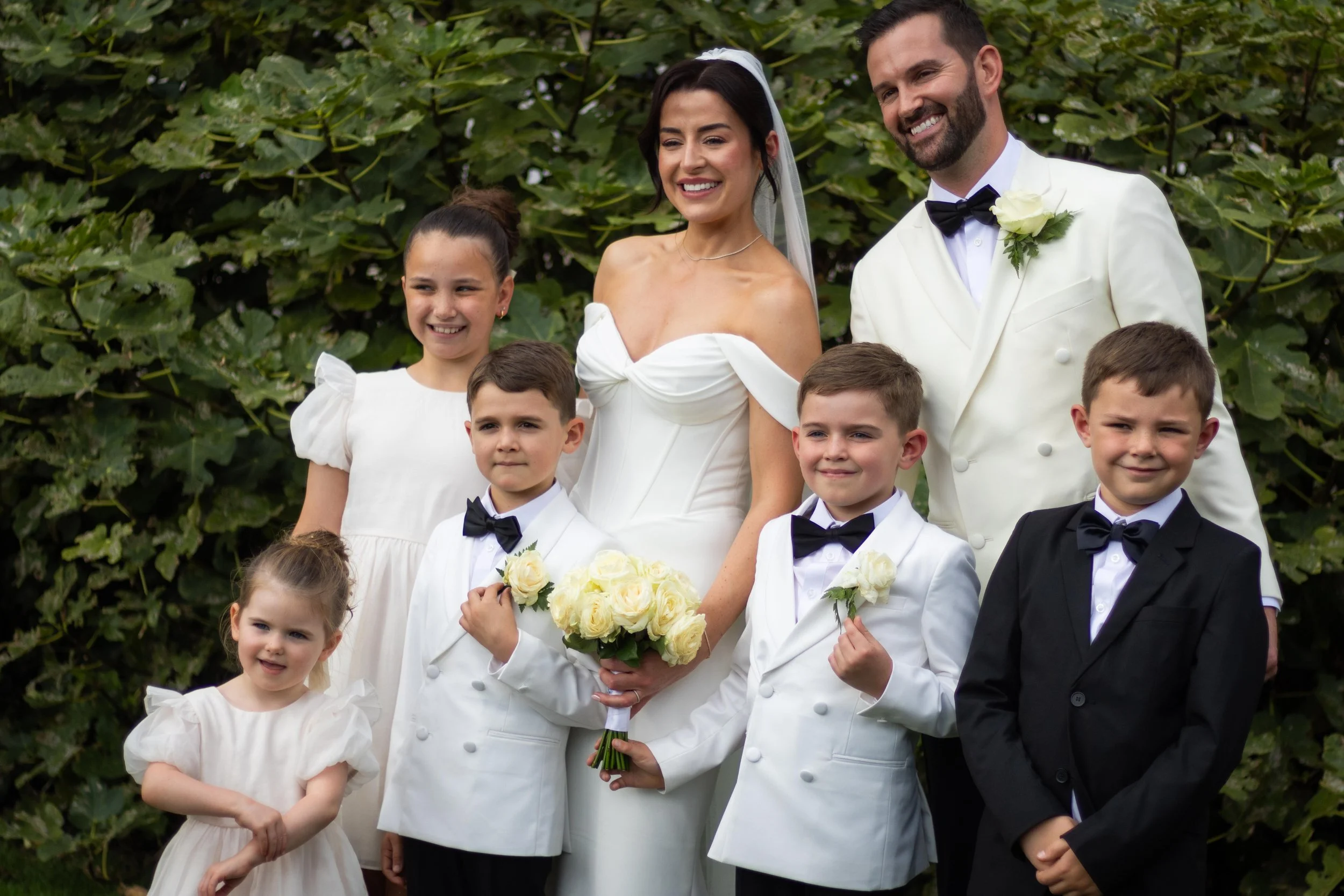 A wedding couple with four children standing outdoors in front of green shrubbery. The bride is holding a bouquet of white roses, and the groom is dressed in a white tuxedo with a black bow tie. The children are wearing formal attire: some in white t