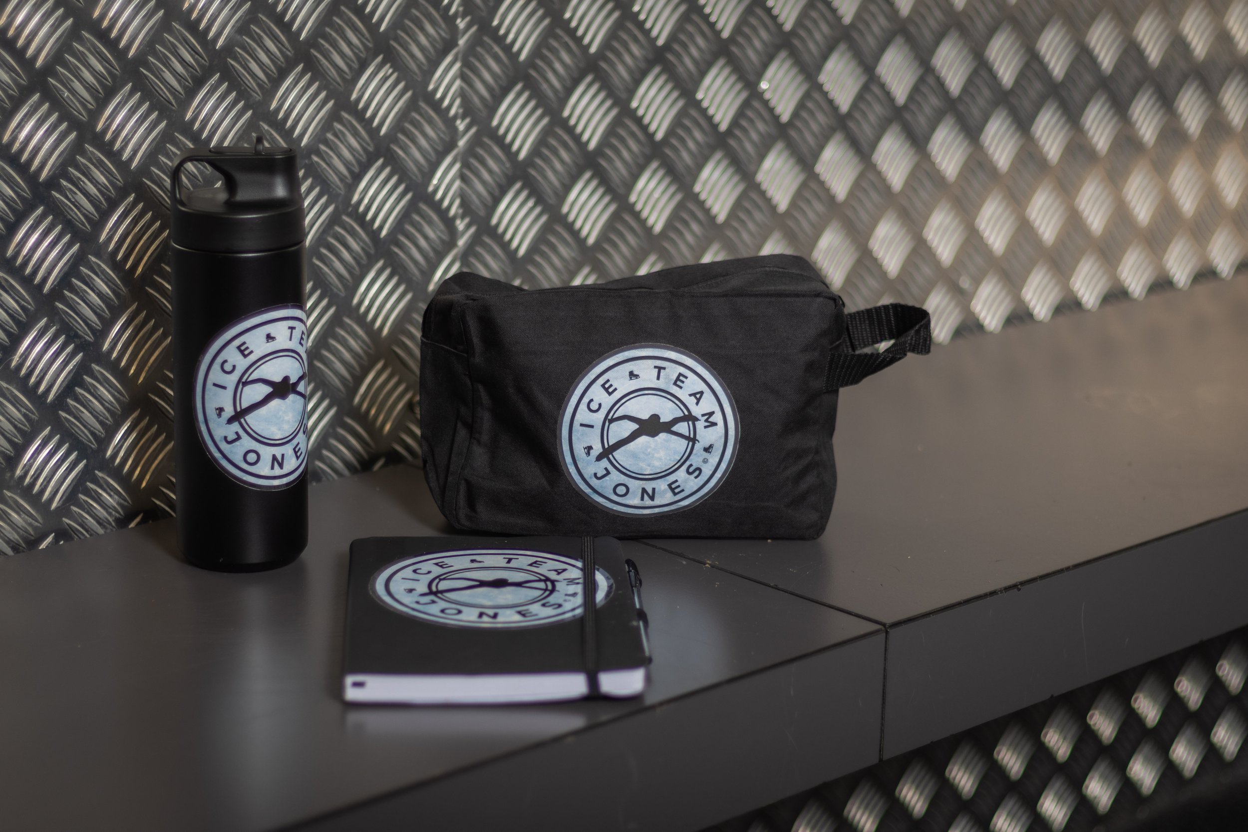 A black travel mug, a black zippered pouch, and a black notebook with an ice and tea logo on a black table against a textured metallic wall.