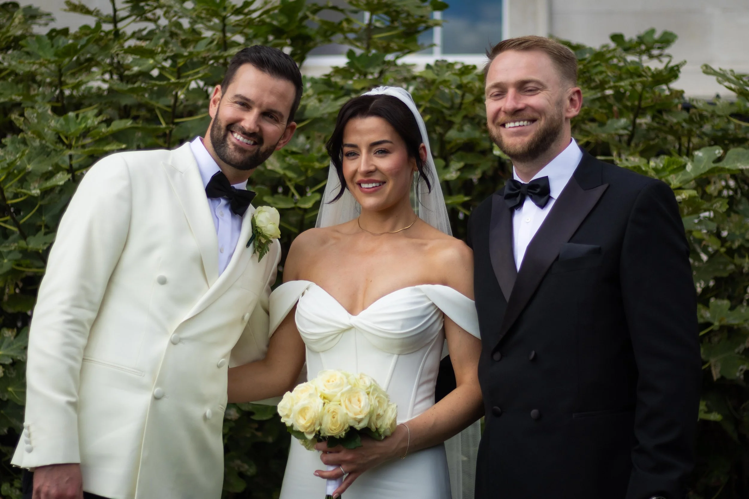 Three people smiling at a wedding: a bride in a white off-shoulder gown holding a bouquet of white roses, and two grooms in tuxedos, one in a white tuxedo and the other in a black tuxedo, standing outdoors with green foliage in the background.