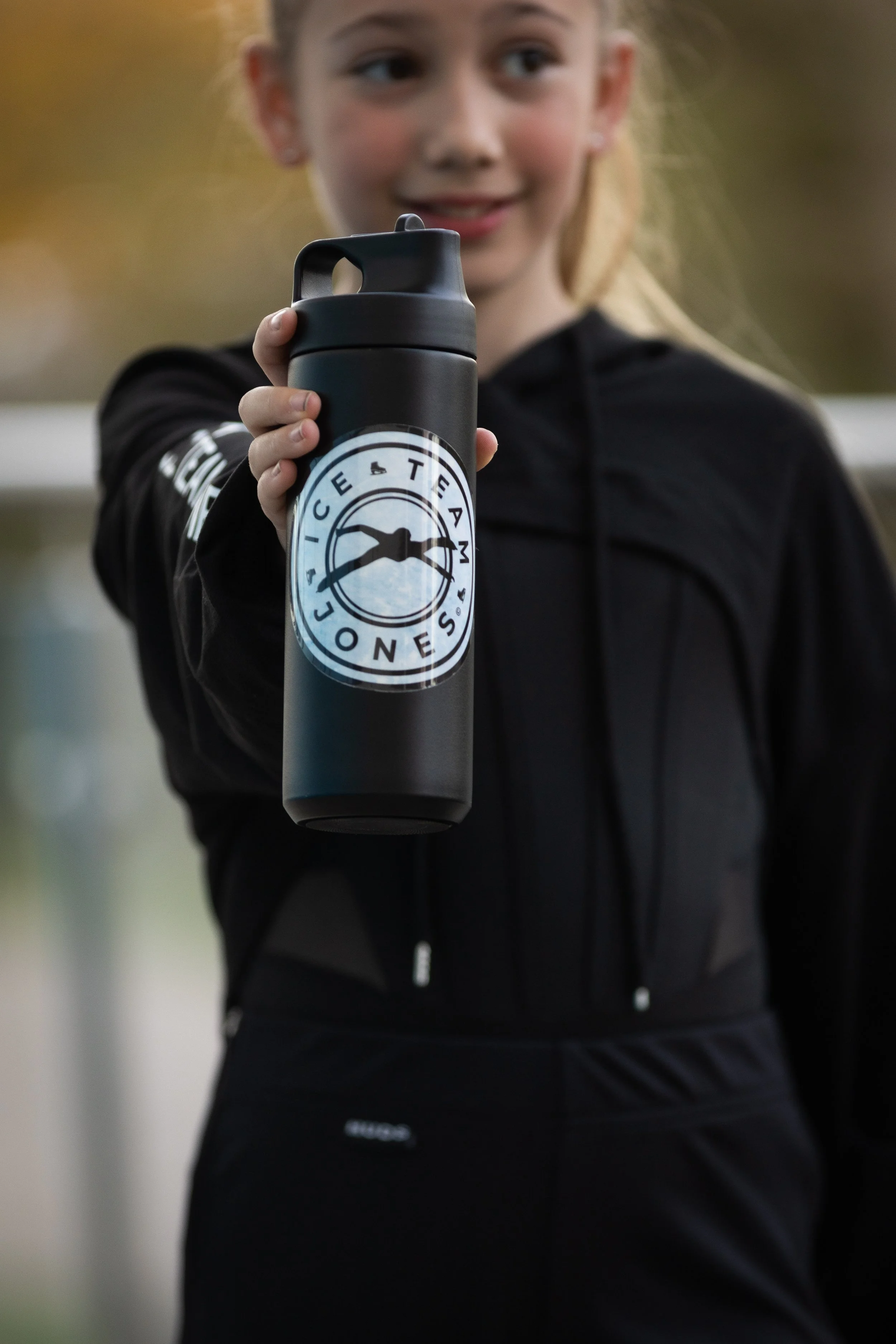 A young girl in athletic clothing holding a black water bottle with a logo that reads 'Ice Team Los Angeles' and features an eye icon.
