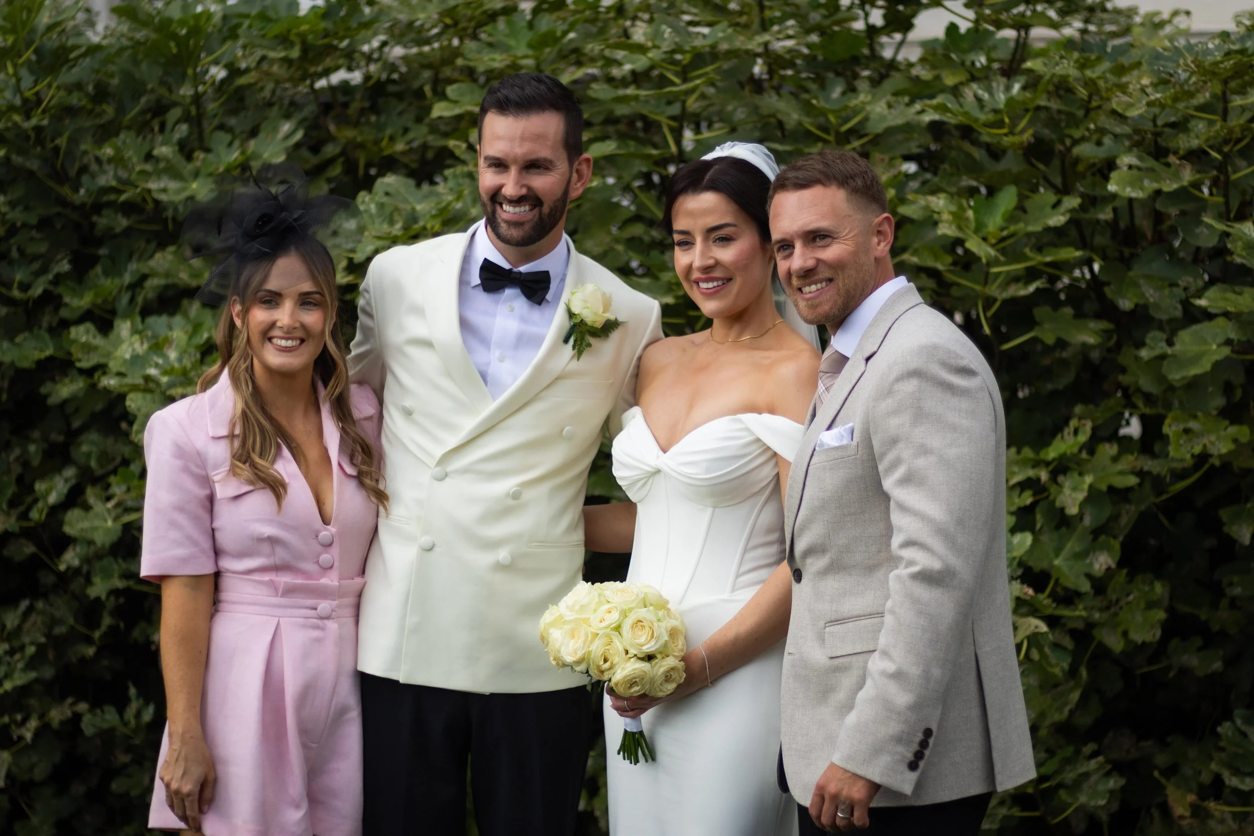 Group of four people at a wedding, standing outdoors in front of greenery, smiling for the camera. The bride in a white dress holding a bouquet of white roses, the groom in a white tuxedo jacket with a black bow tie, and two other people in pastel su