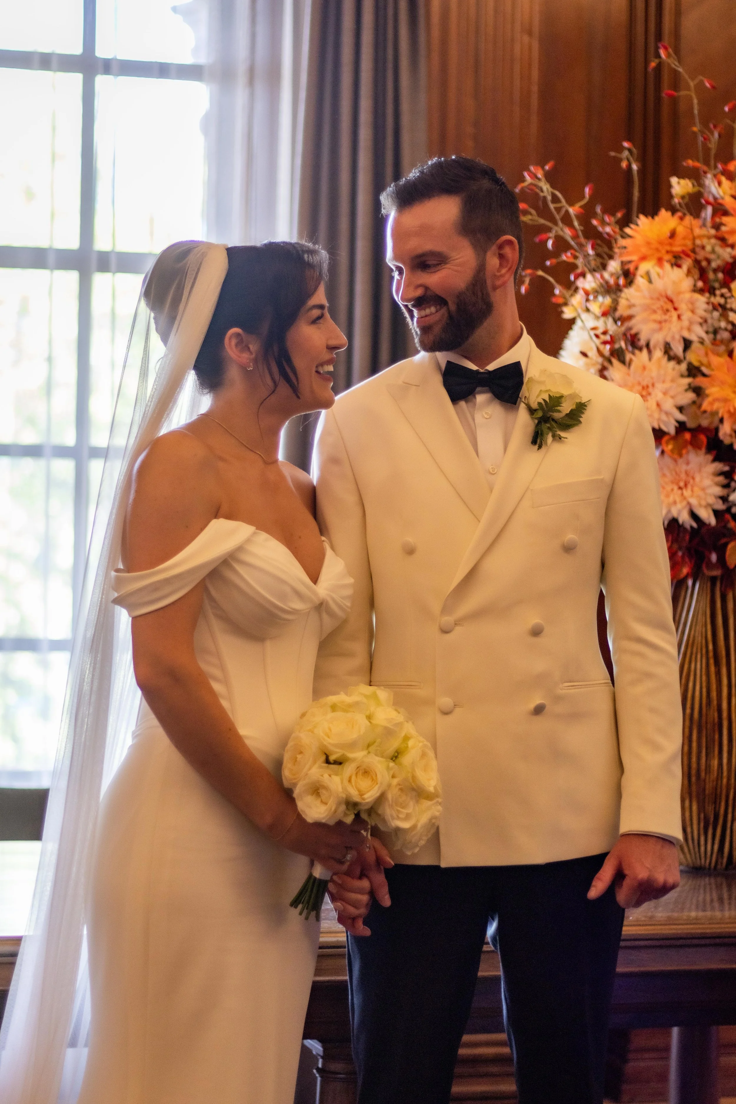 A bride and groom on their wedding day, smiling at each other indoors with a large bouquet of white roses and a floral arrangement in the background.