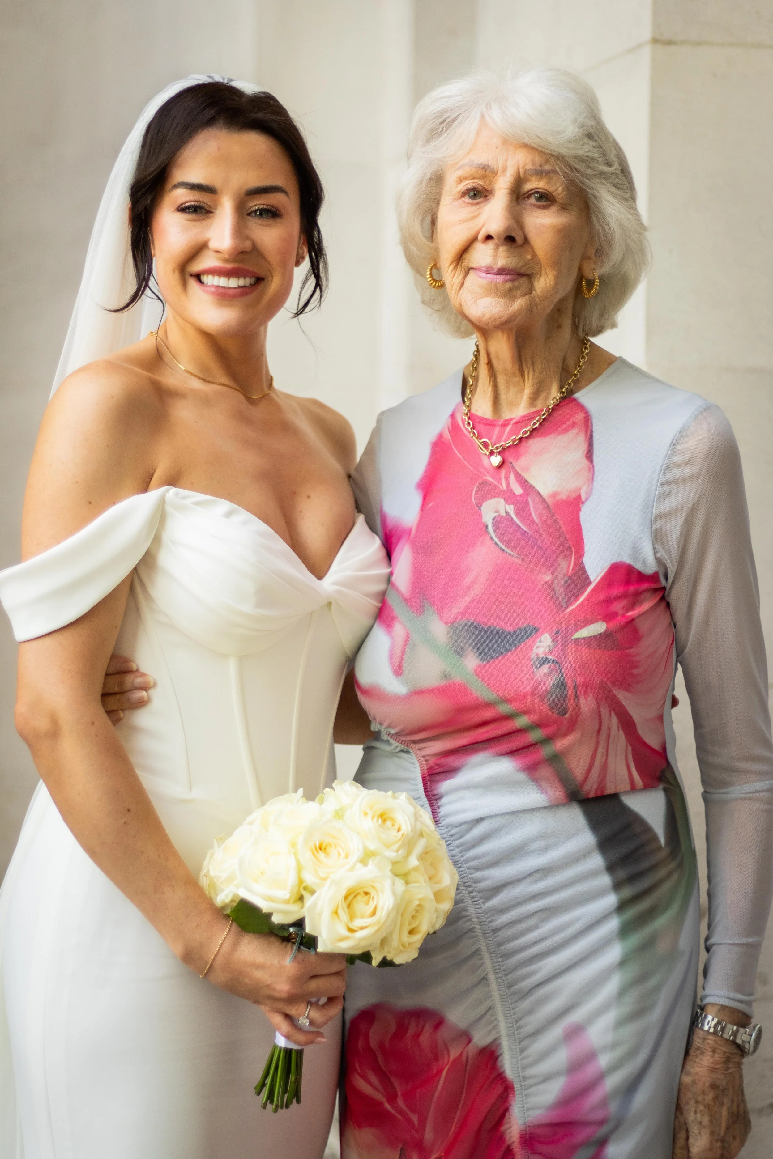 A bride in a white wedding dress holding a bouquet of white roses, standing next to an elderly woman in a floral dress, both smiling at the camera.