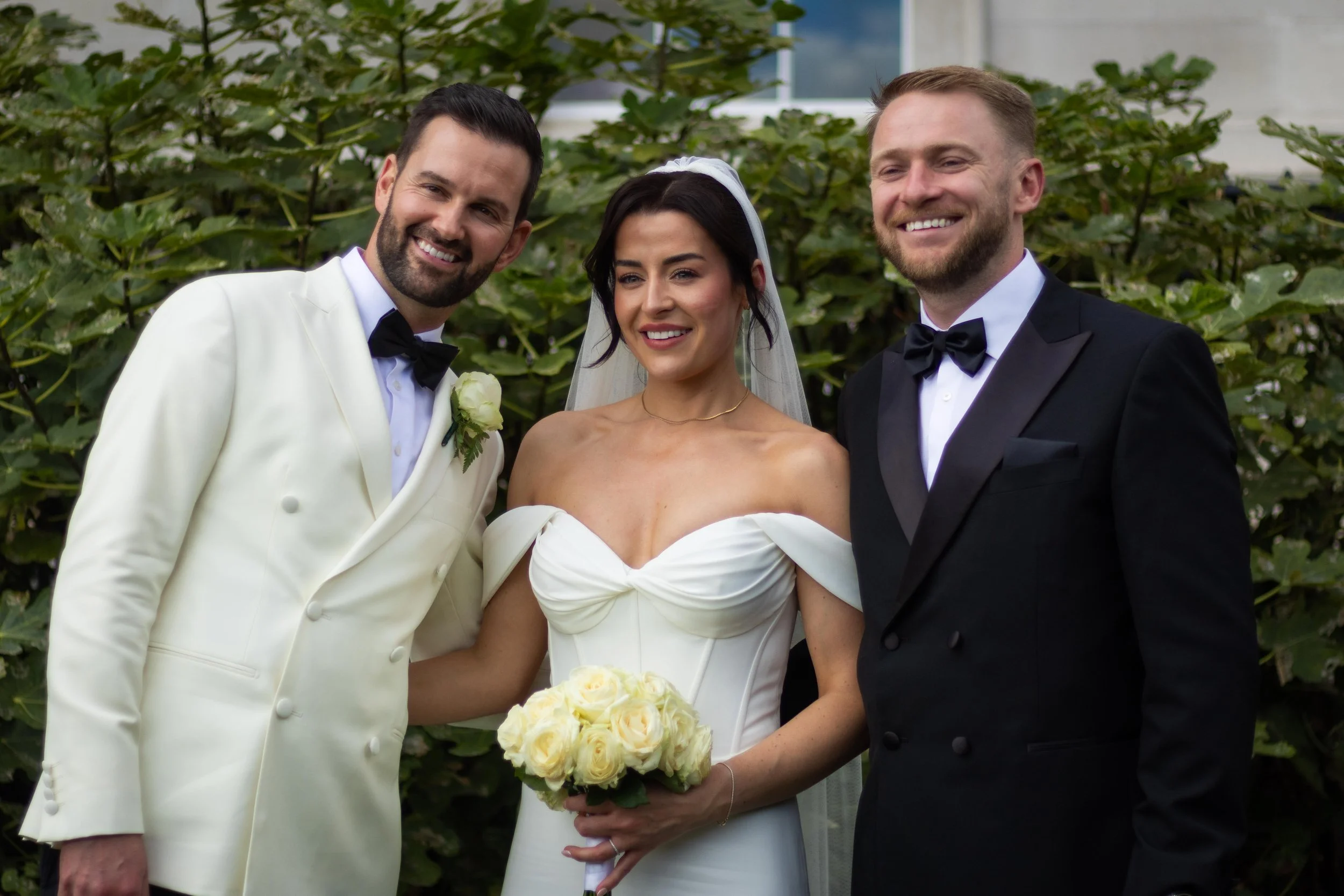 A bride in a white wedding dress holding a bouquet of white roses, standing between two grooms in tuxedos, outdoors with greenery in the background.