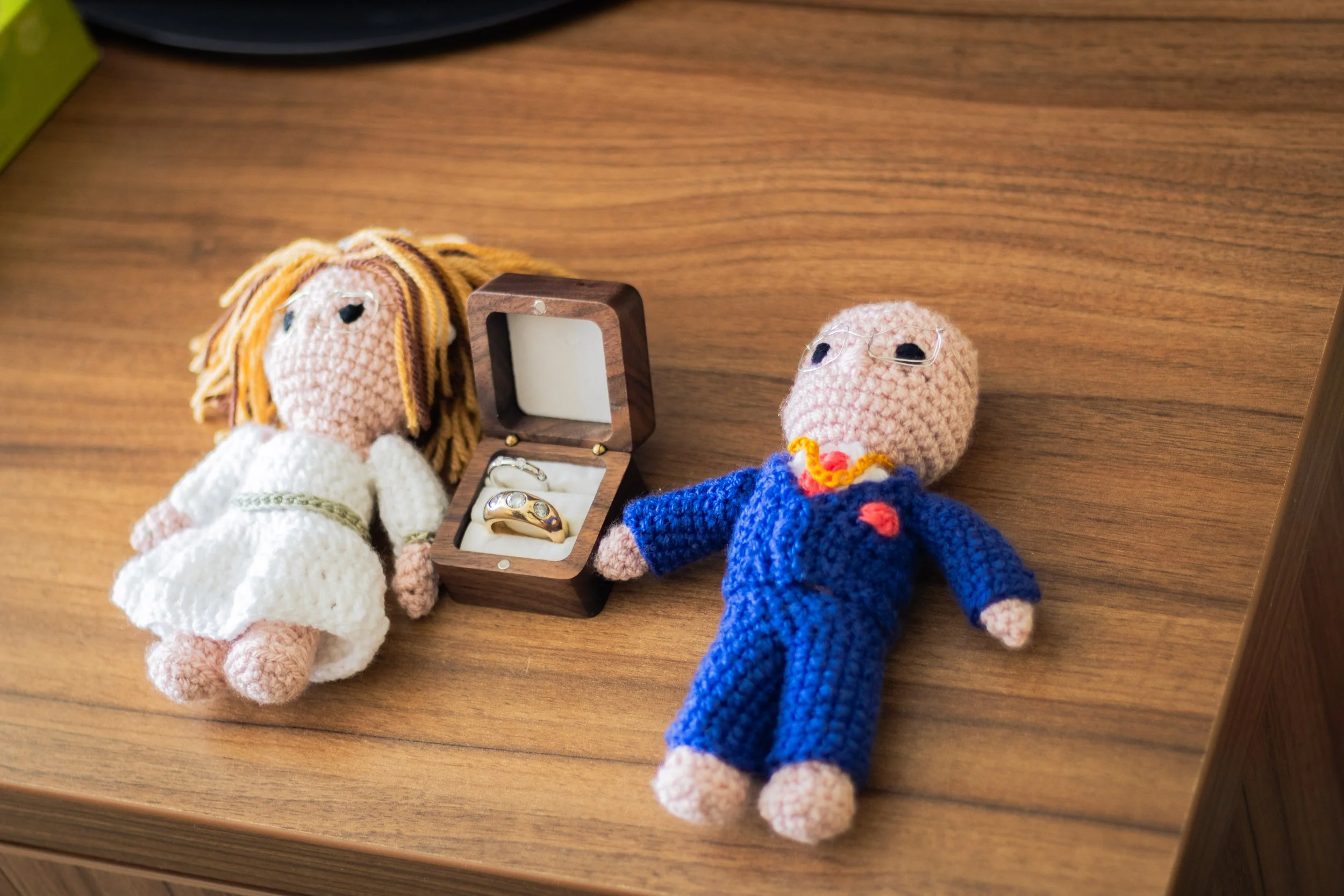 Two crocheted dolls, one dressed as a bride and the other as a groom, are lying on a wooden surface. The bride doll has yellow yarn hair, a white dress, and the groom doll has glasses, a blue suit, and a red bow tie. Between them is a small open wood
