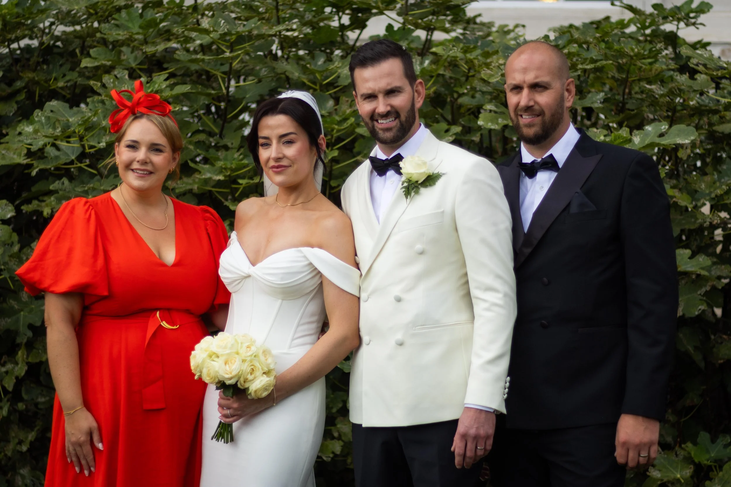 A group photograph of five people in formal attire at a wedding, standing outdoors with green foliage in the background. The bride holds a bouquet of white roses, and a woman in a red dress stands on her left. Two men in tuxedos, one with a white tux
