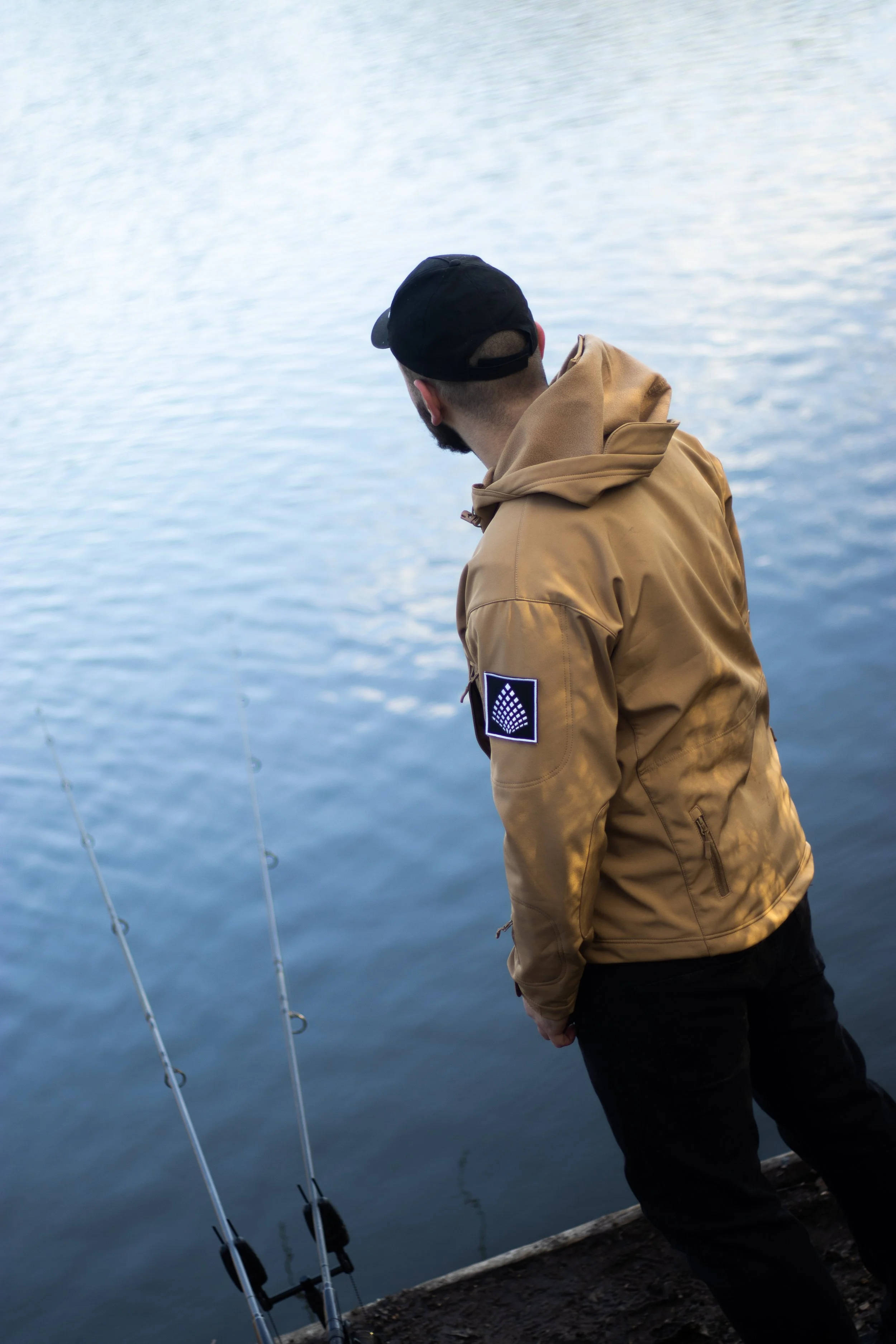 Man wearing a tan jacket and black cap standing on a dock, fishing by the water with two fishing rods.