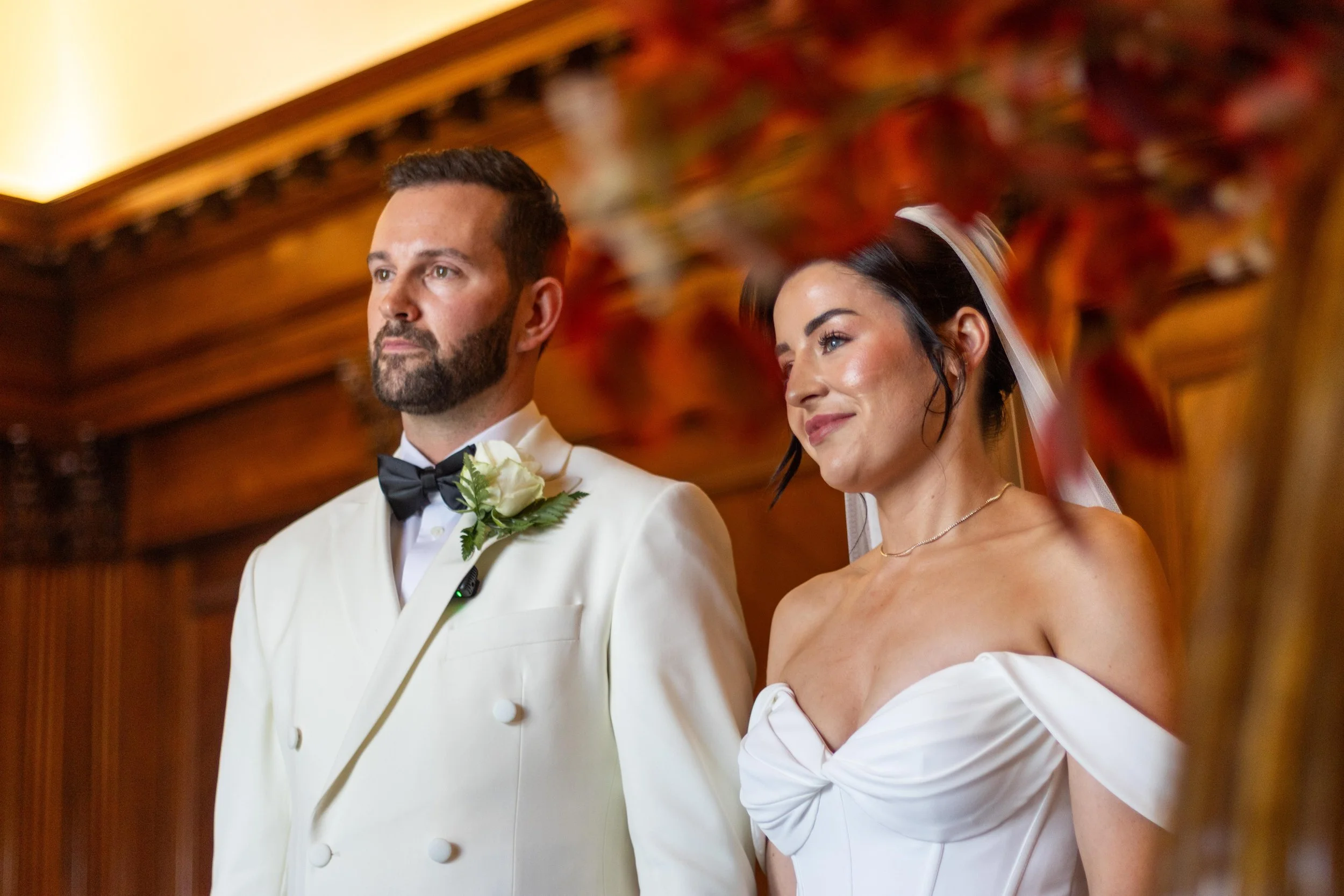 A bride and groom standing at their wedding ceremony, with the groom wearing a white tuxedo with a black bow tie and boutonniere, and the bride in a strapless white wedding dress with a veil, in a wood-paneled room.