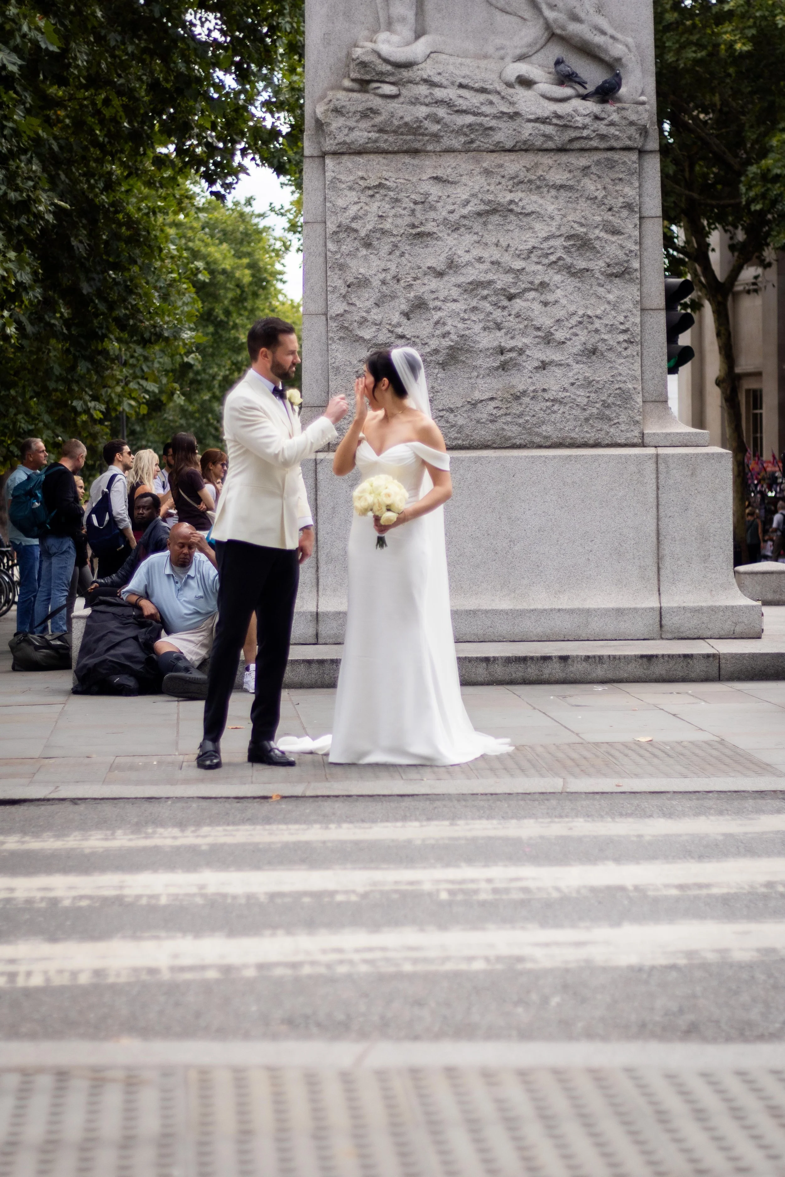 A wedding ceremony taking place outdoors in front of a stone monument with a bride and groom exchanging vows. The bride, in a white gown holding a bouquet, and the groom, in a white tuxedo jacket, are facing each other. A crowd of spectators is watch