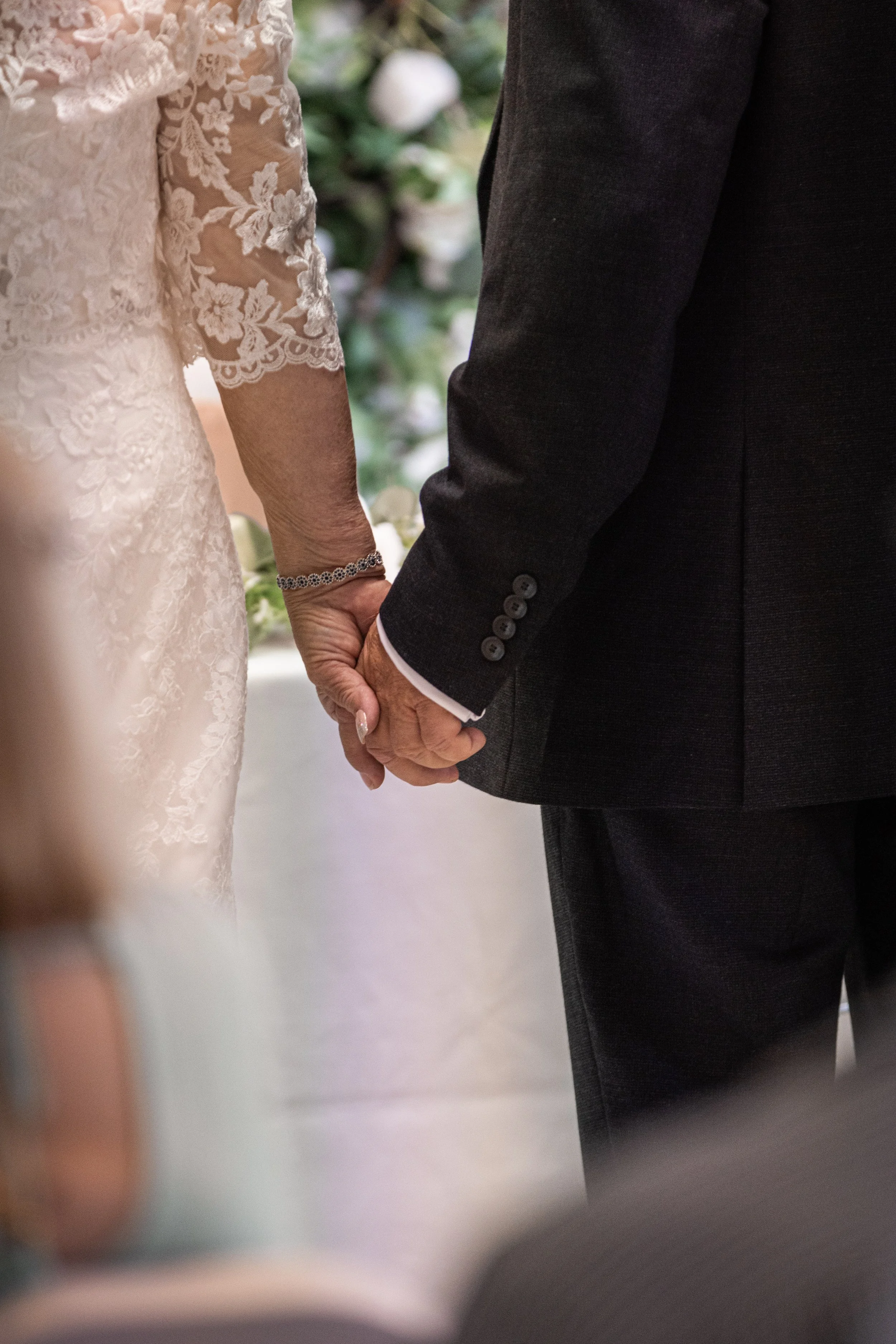 A close-up of an elderly couple holding hands during a wedding ceremony, with the woman wearing a white lace dress and a bracelet, and the man in a dark suit.