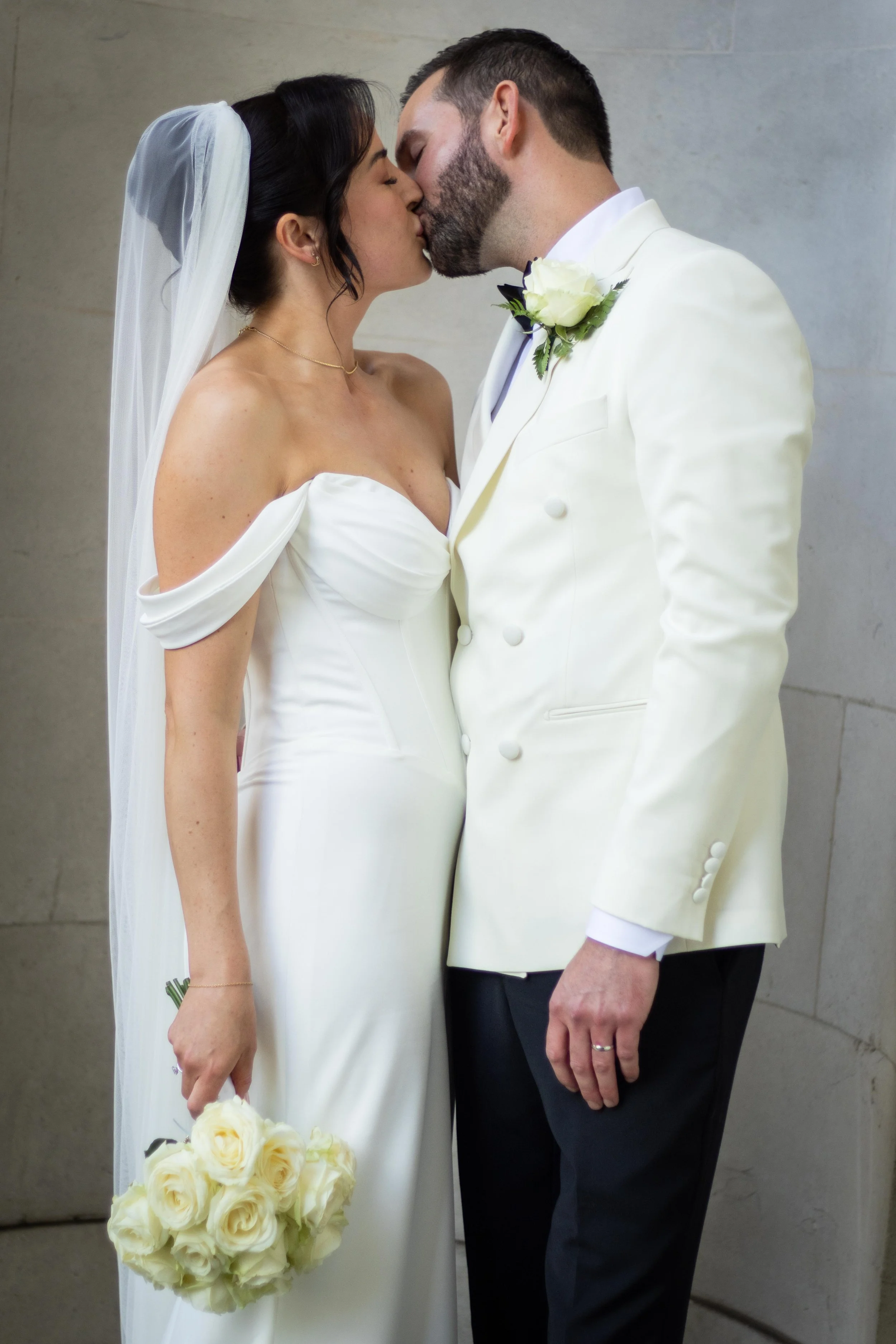 A bride and groom kissing, dressed in wedding attire, with the bride holding a bouquet of white roses.