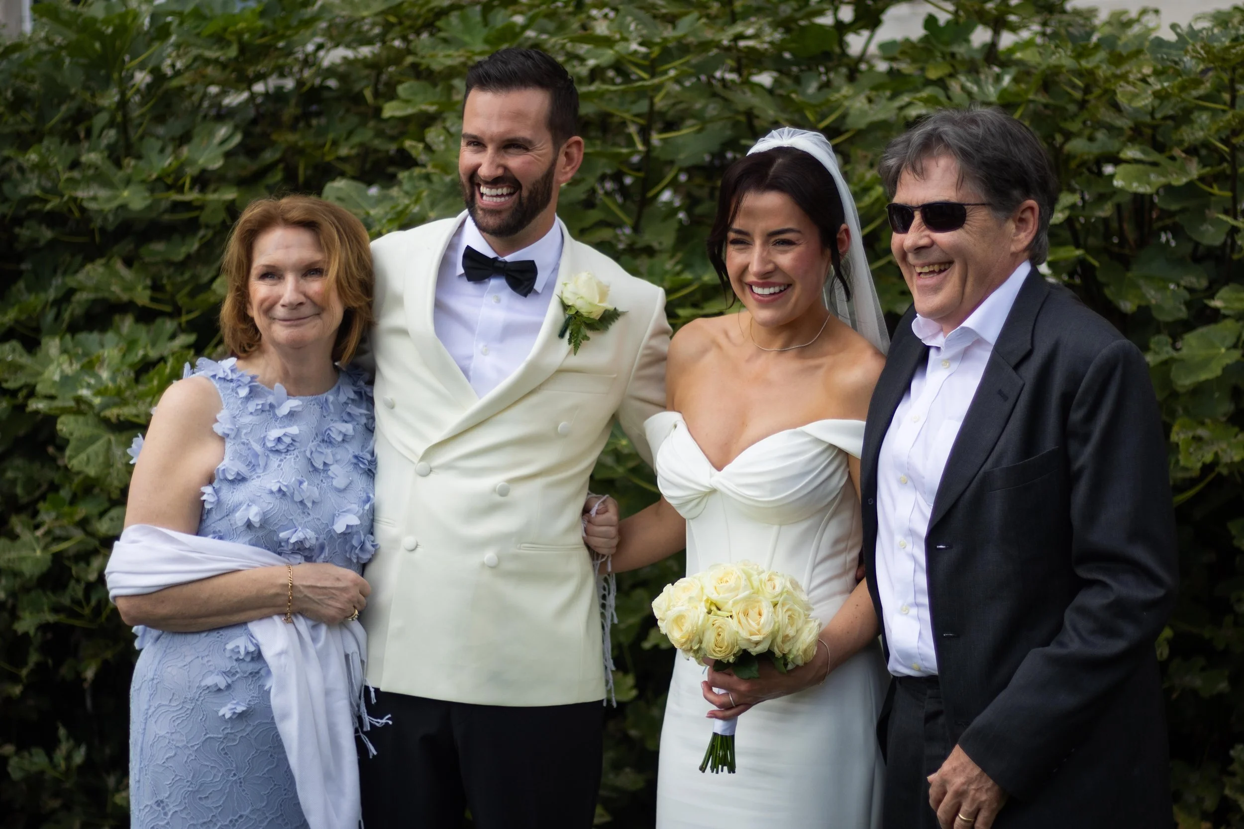 Group of four people celebrating at a wedding, standing outdoors with green foliage in the background. A bride in a white gown holding a bouquet of yellow roses, a groom in a cream tuxedo, and two older adults, a woman in a light blue dress with flor