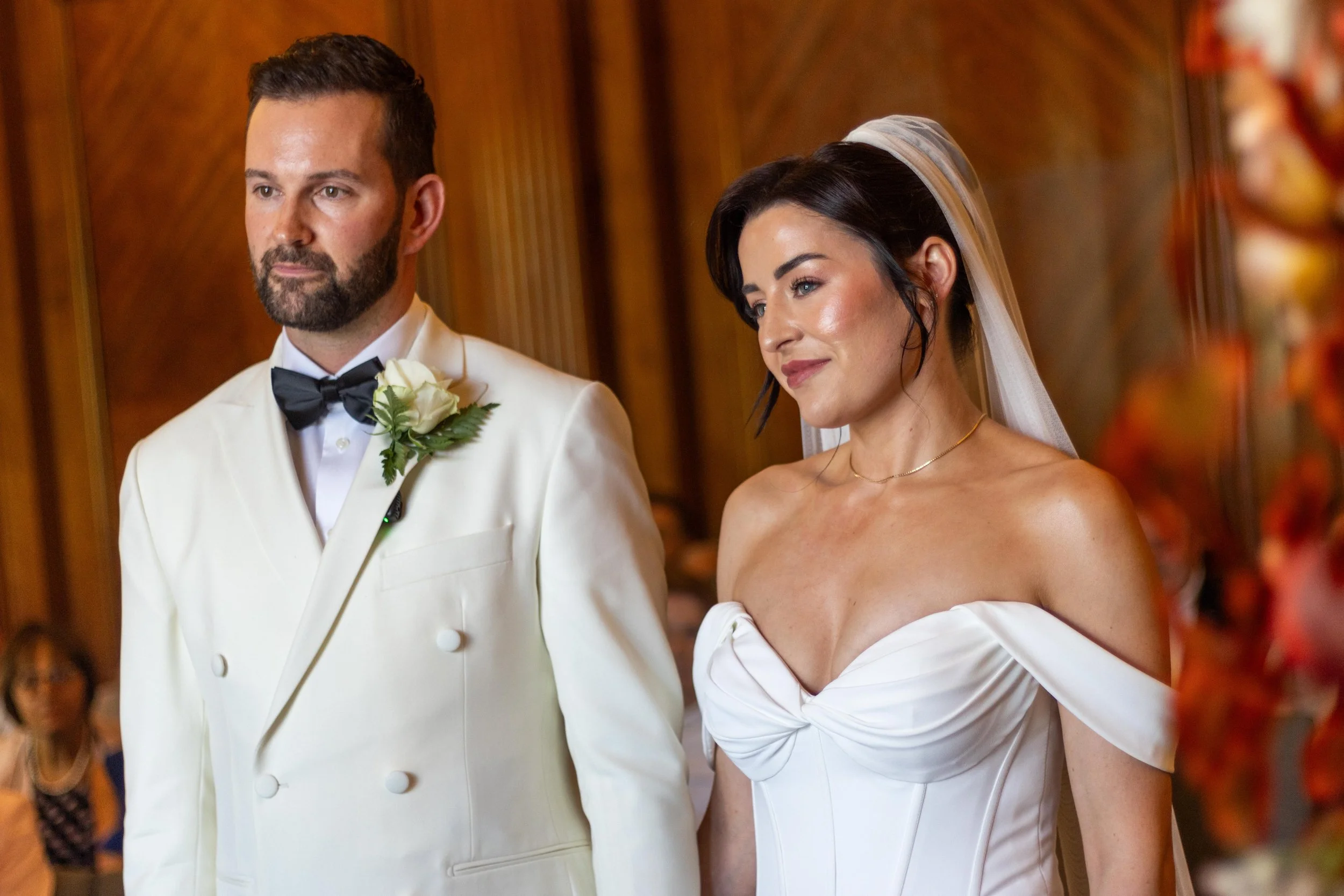 A bride and groom standing during their wedding ceremony, with the groom in a white tuxedo and black bow tie, and the bride in a white off-the-shoulder wedding gown and veil.