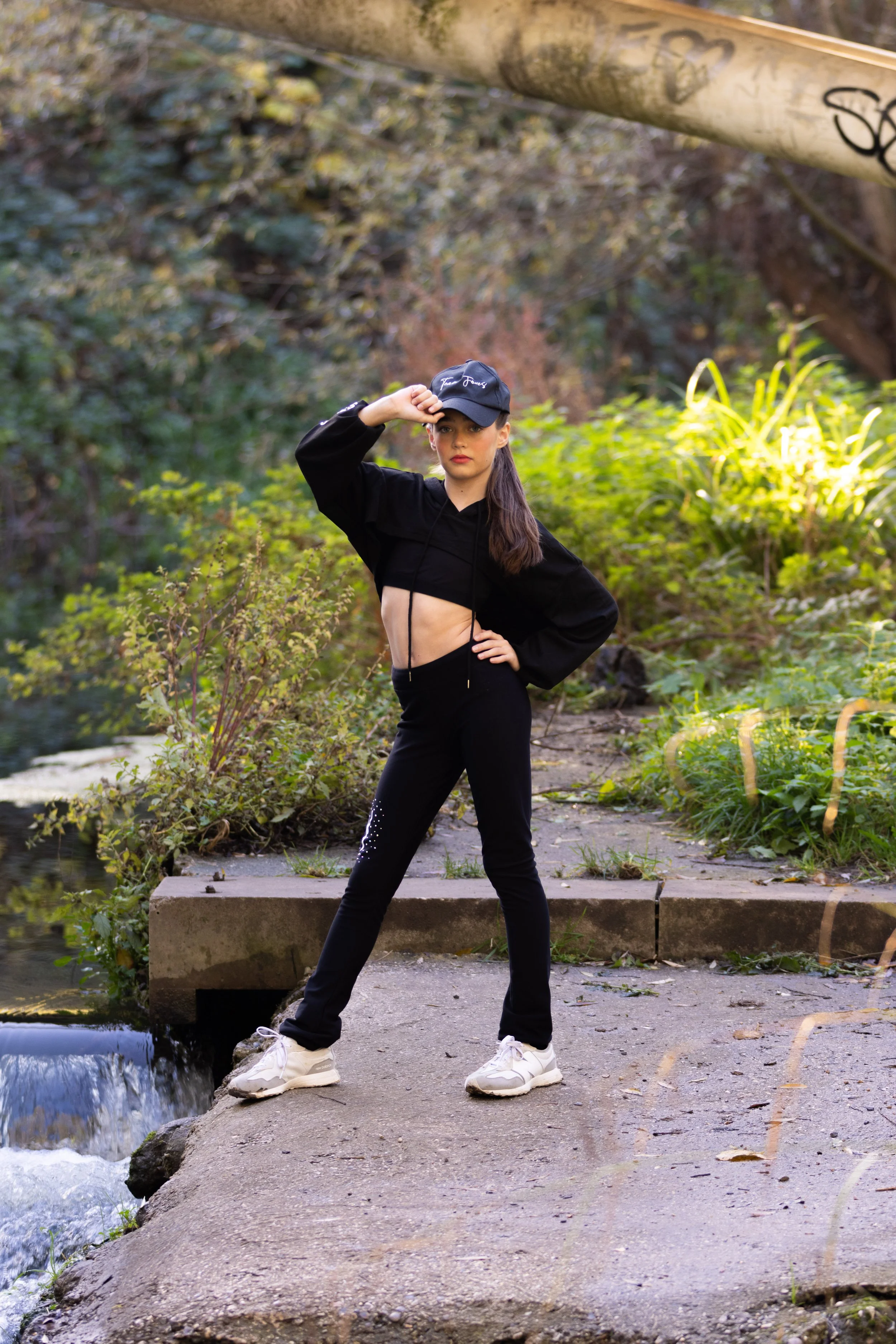 A young woman standing on a concrete path by a small stream in a lush, green outdoor park, wearing a black hoodie, black pants with rhinestones, white sneakers, and a black cap, with one hand on her hip and the other adjusting her cap.