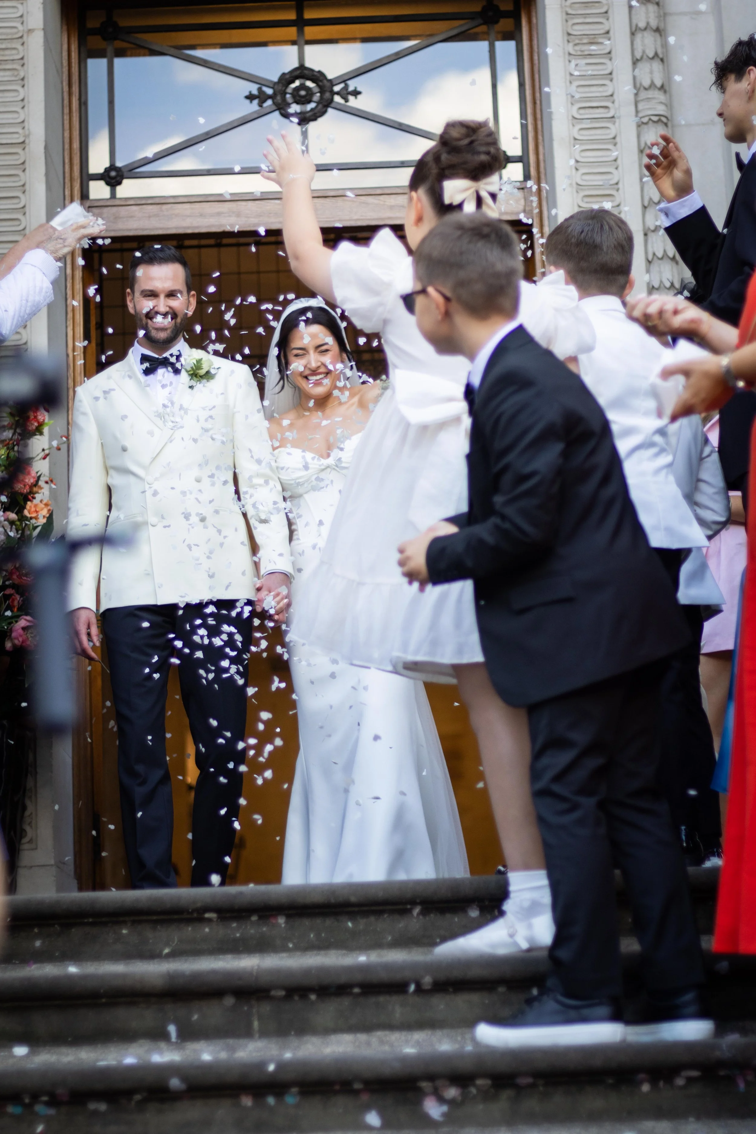 A newlywed couple exiting a building, holding hands and smiling, while guests throw confetti around them during a celebration.