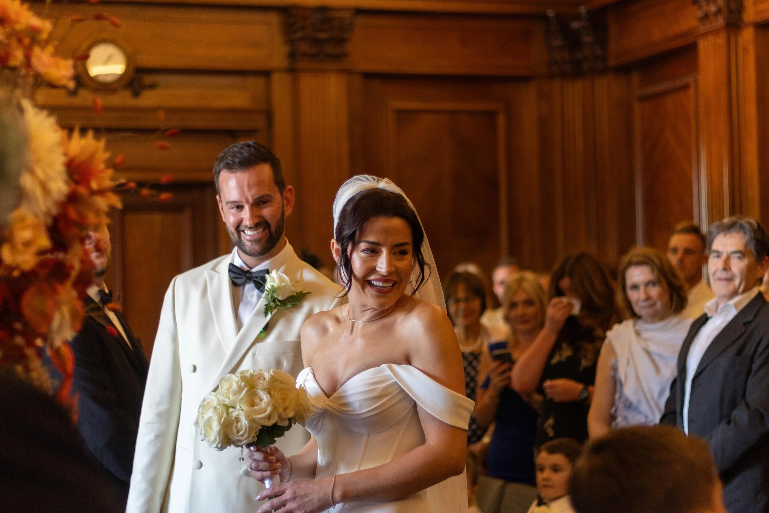 Bride and groom at wedding ceremony, surrounded by guests in a wood-paneled room.