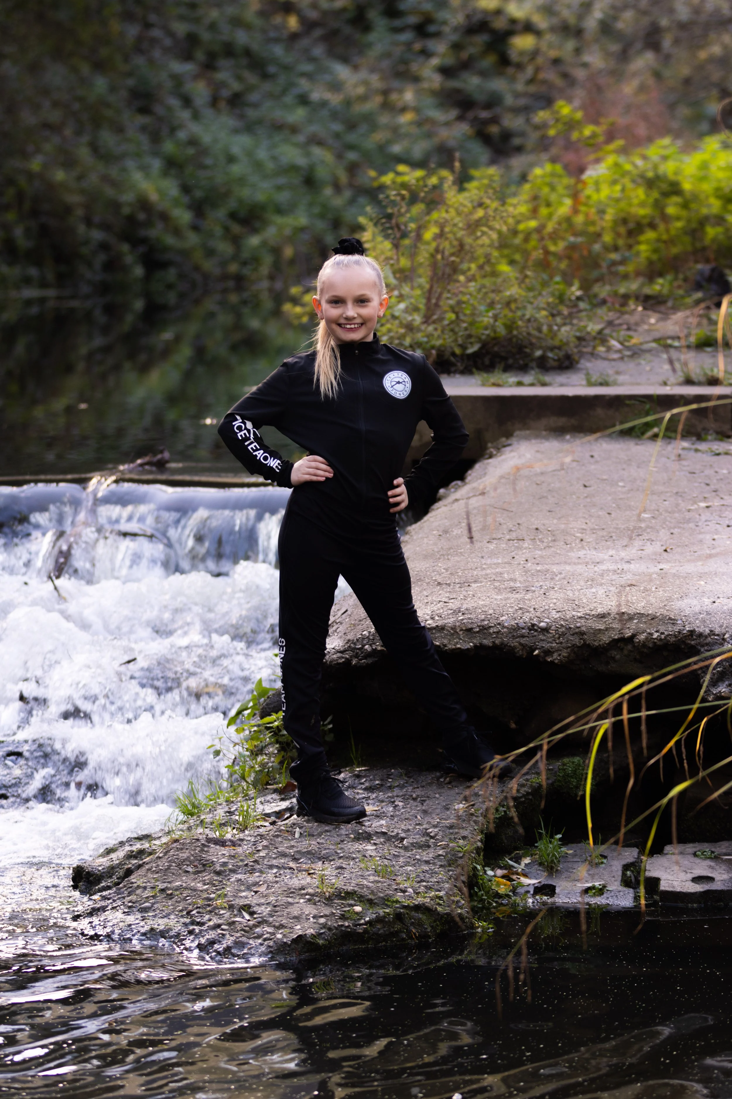 A young girl stands confidently on a rock near a flowing stream in a forest, smiling at the camera, wearing a black athletic tracksuit.