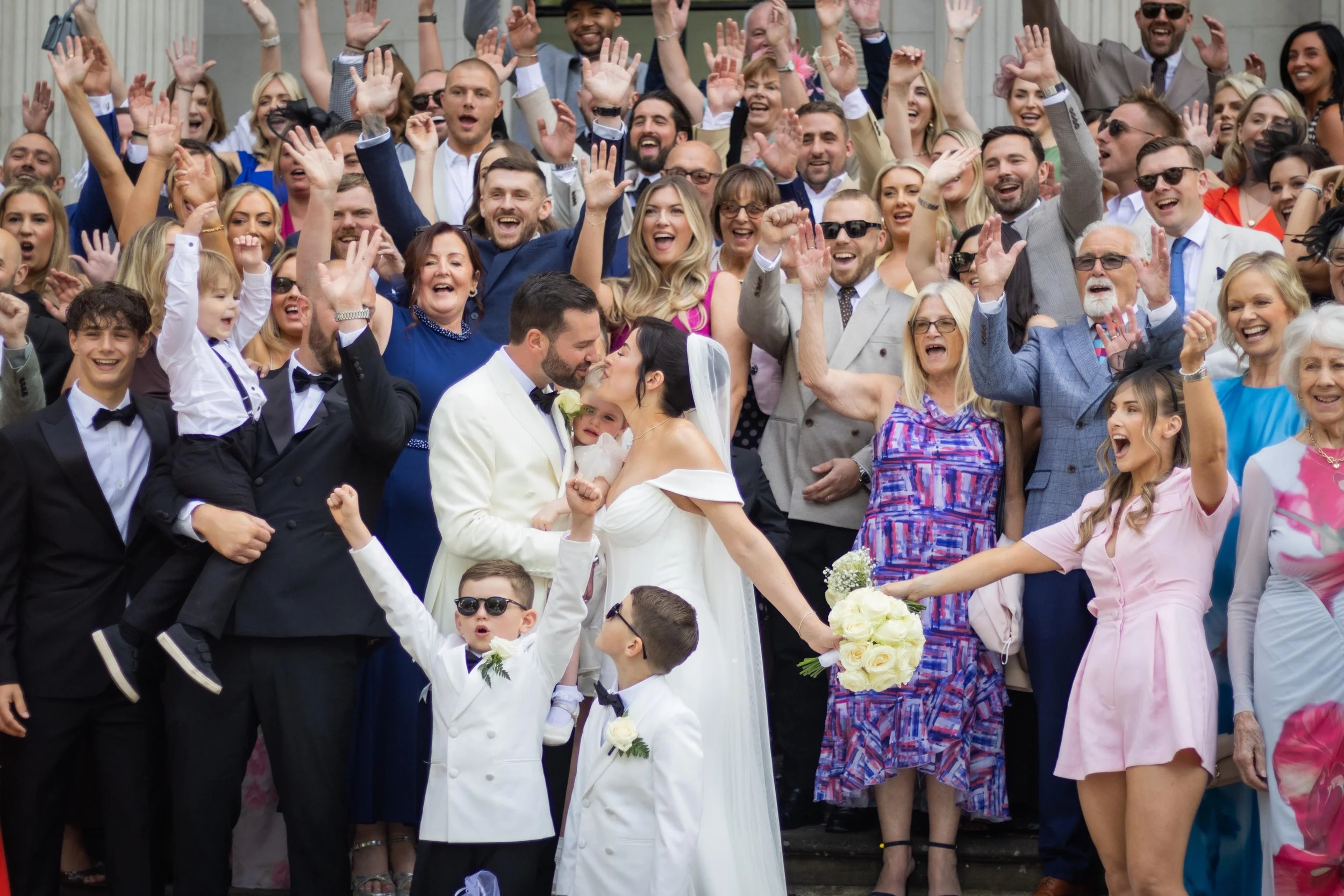 A large group of people at a wedding, with the bride and groom in the center kissing. The crowd is smiling, cheering, raising their hands, and celebrating. The bride is holding a bouquet of white roses.