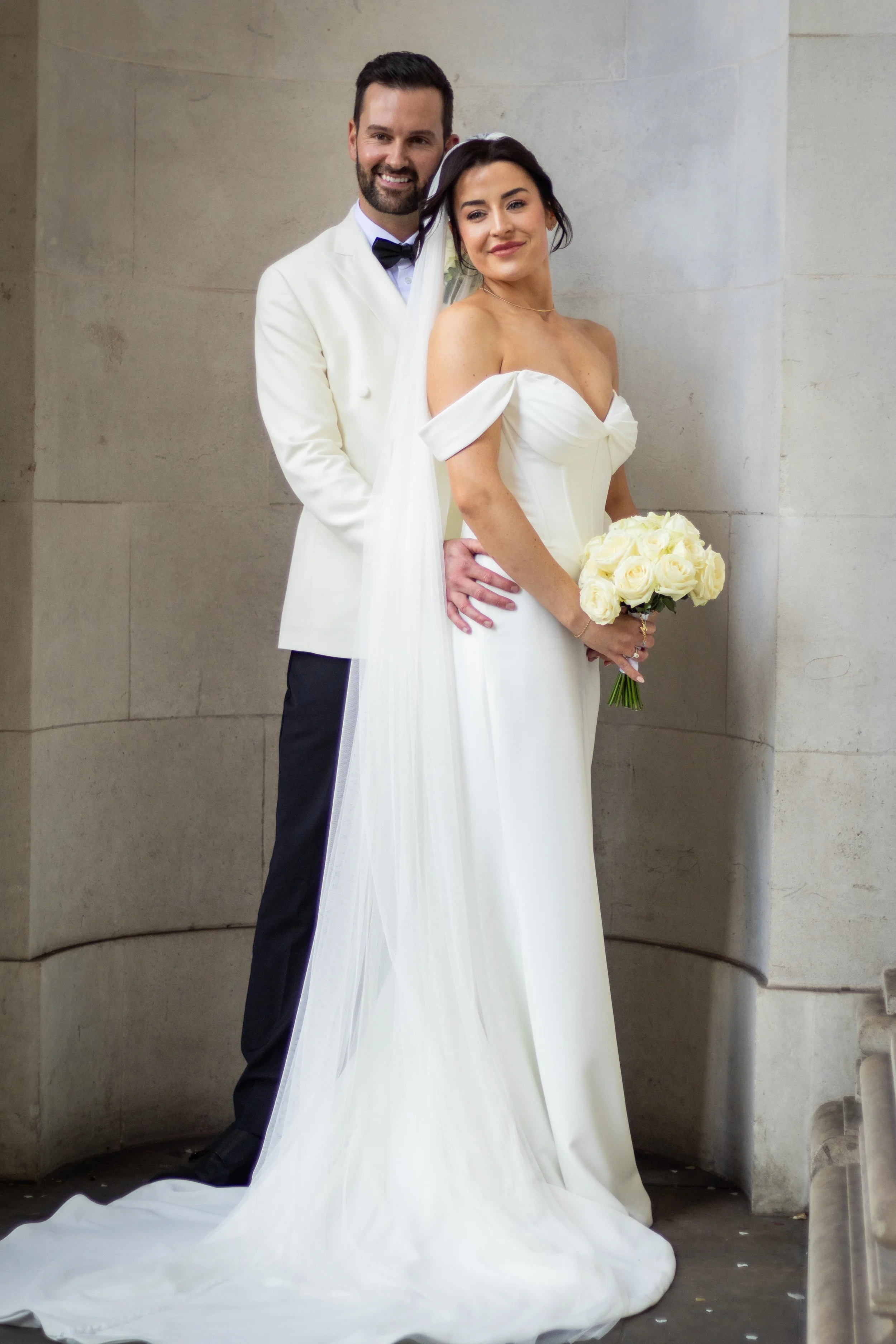 A bride and groom dressed in wedding attire standing together against a stone wall, with the bride holding a bouquet of white roses.