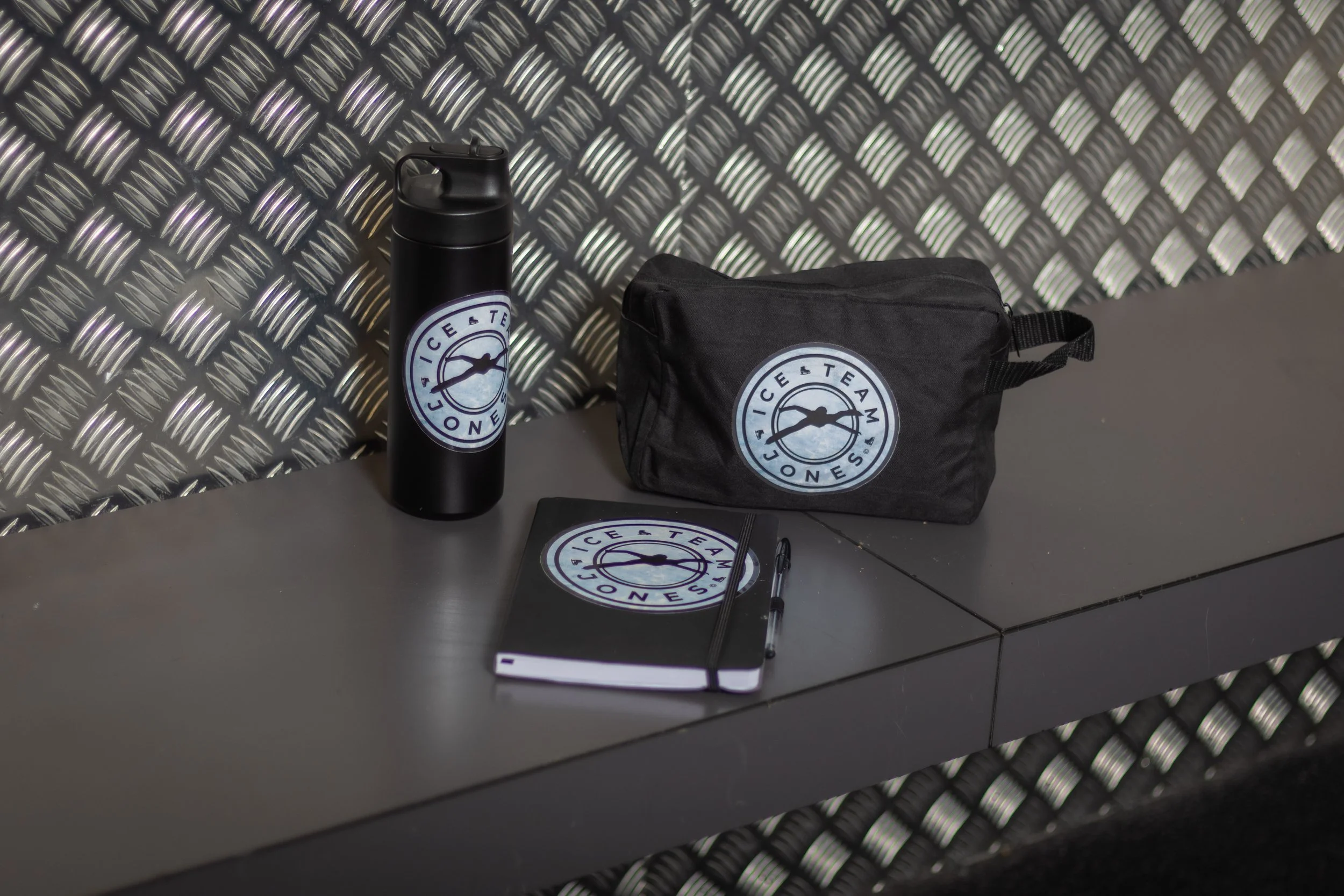 A black water bottle, a black zippered pouch, and a black notebook with a pen resting on it, all with a logo that says "Ice & Teami JONES" on a gray table against a textured metal background.