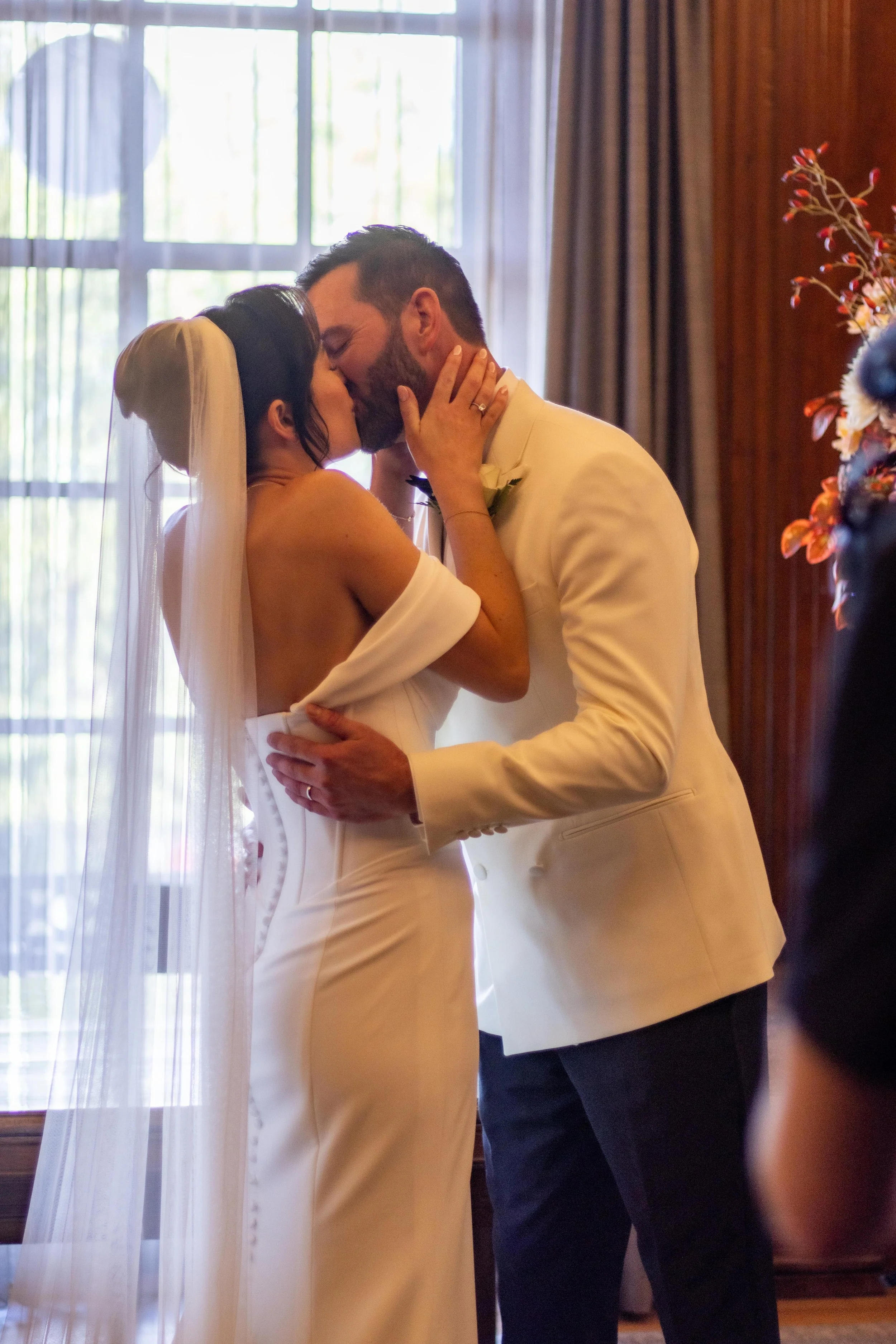 A bride and groom share a kiss during their wedding ceremony indoors, holding each other close.