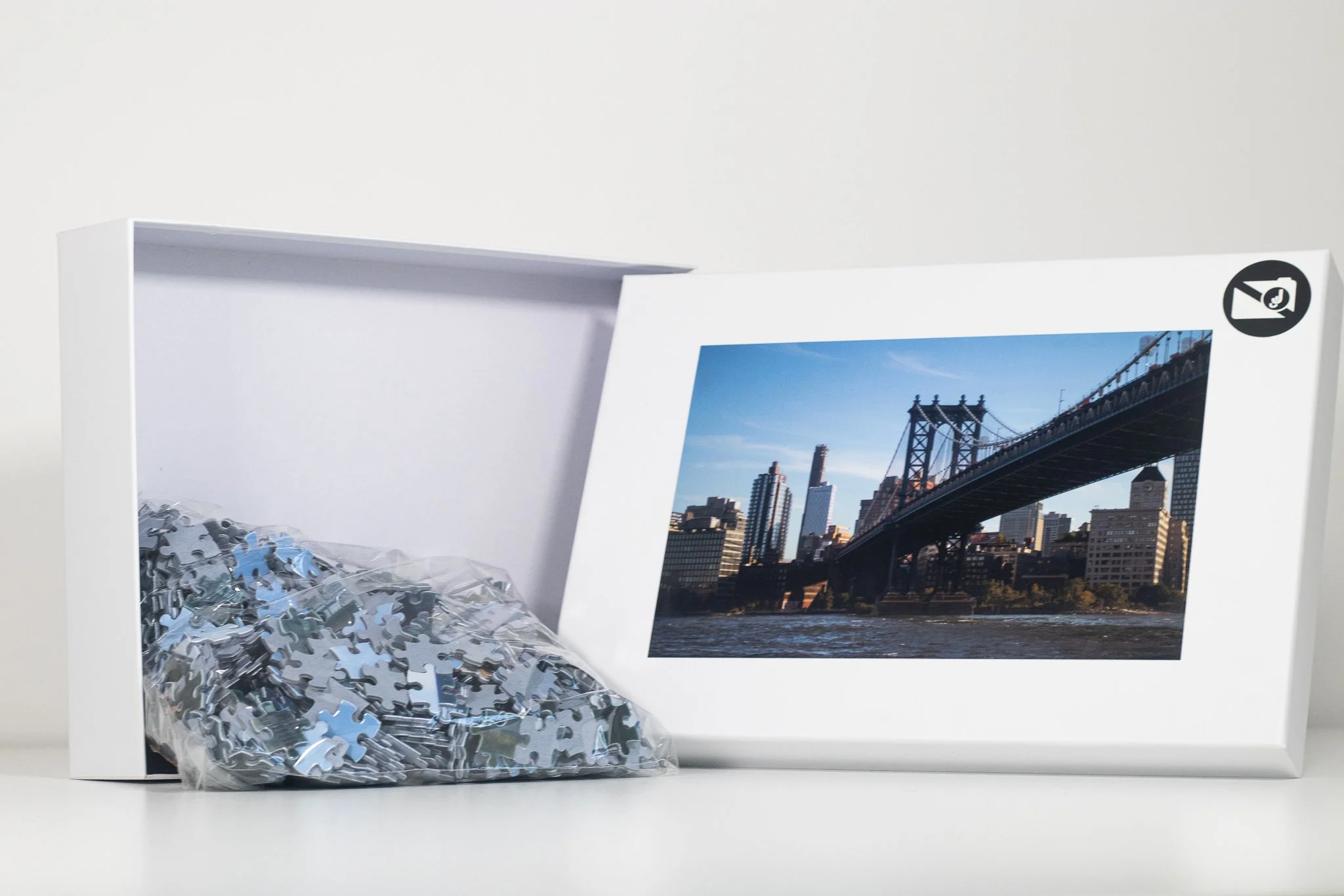 A box with its lid open, containing a pile of blue, silver, and black puzzle pieces, with a photograph of a cityscape and a bridge on the inside of the lid.