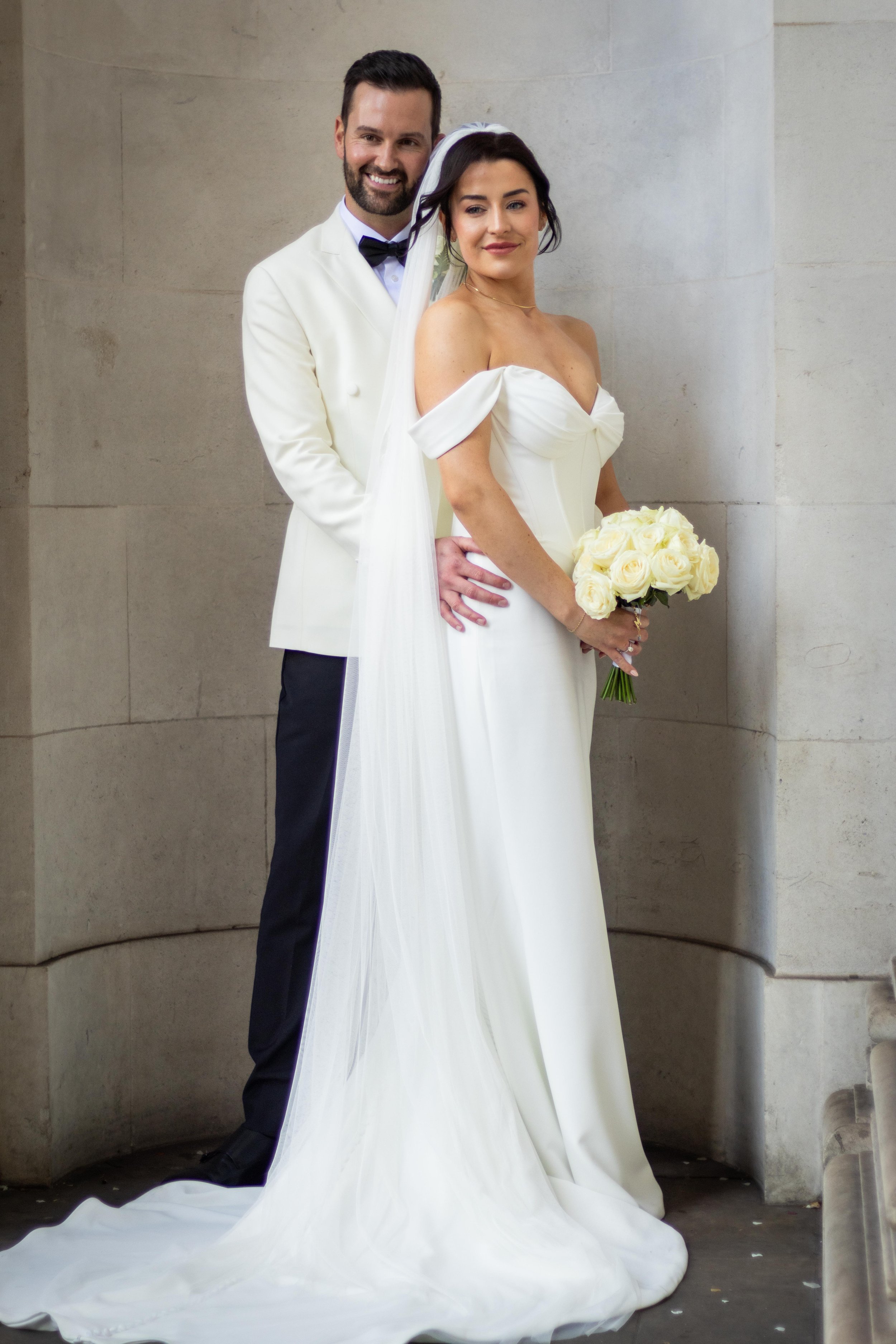 A newlywed couple in wedding attire, standing indoors against a stone wall. The bride holds a bouquet of white roses, and she wears an off-shoulder white wedding gown with a long train. The groom is dressed in a white tuxedo jacket with a black bow t