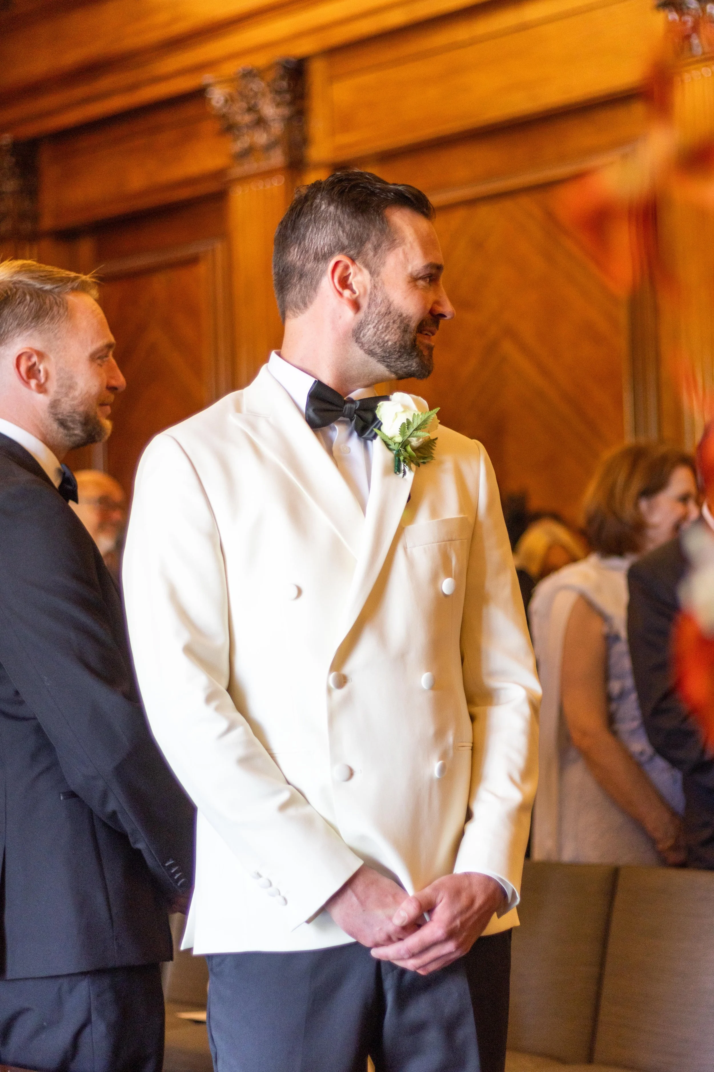 A groom in a white tuxedo jacket with a black bow tie and white flower boutonniere standing during a wedding ceremony, smiling, inside a beautifully decorated wooden hall.