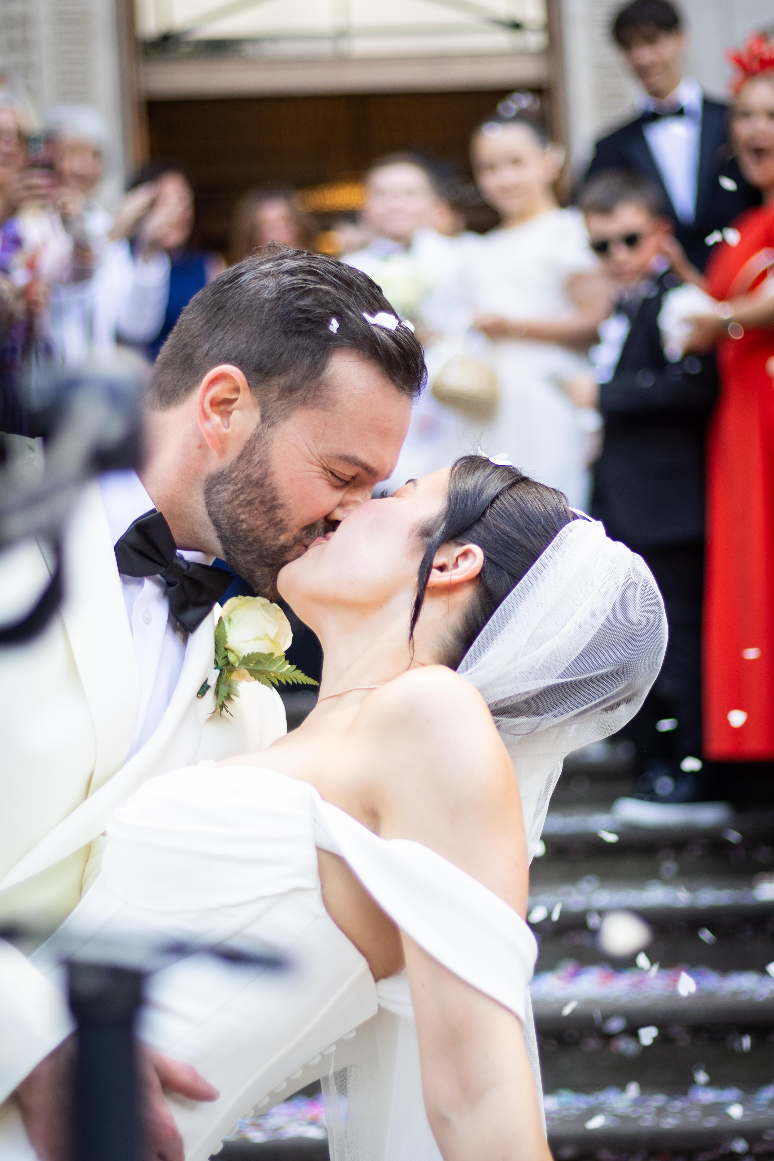 A newlywed couple sharing a kiss at their wedding with guests in the background celebrating.