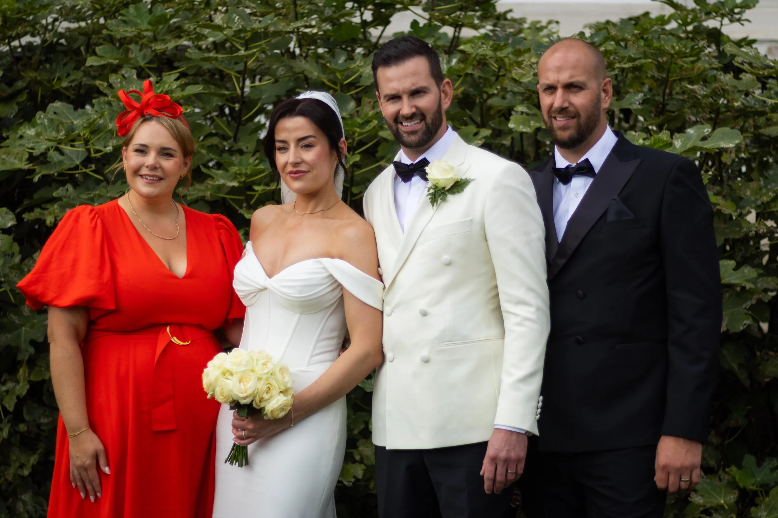 A group of five people dressed in wedding attire outdoors, surrounded by greenery, including a bride holding a bouquet of white roses, a groom in a white tuxedo, and three guests in elegant dresses and suits.