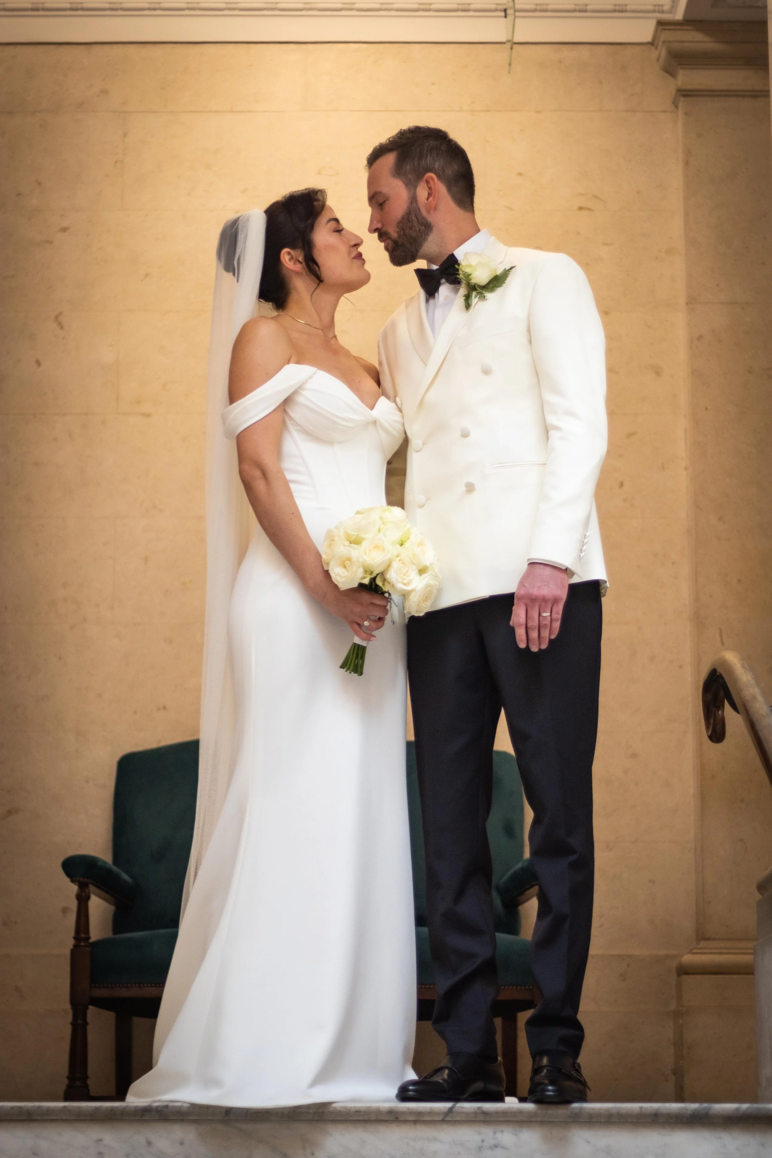 A bride and groom standing close together, about to kiss during their wedding ceremony indoors. The bride is in a white off-the-shoulder gown holding a bouquet of white roses, and the groom is in a white tuxedo jacket with black pants, a black bow ti