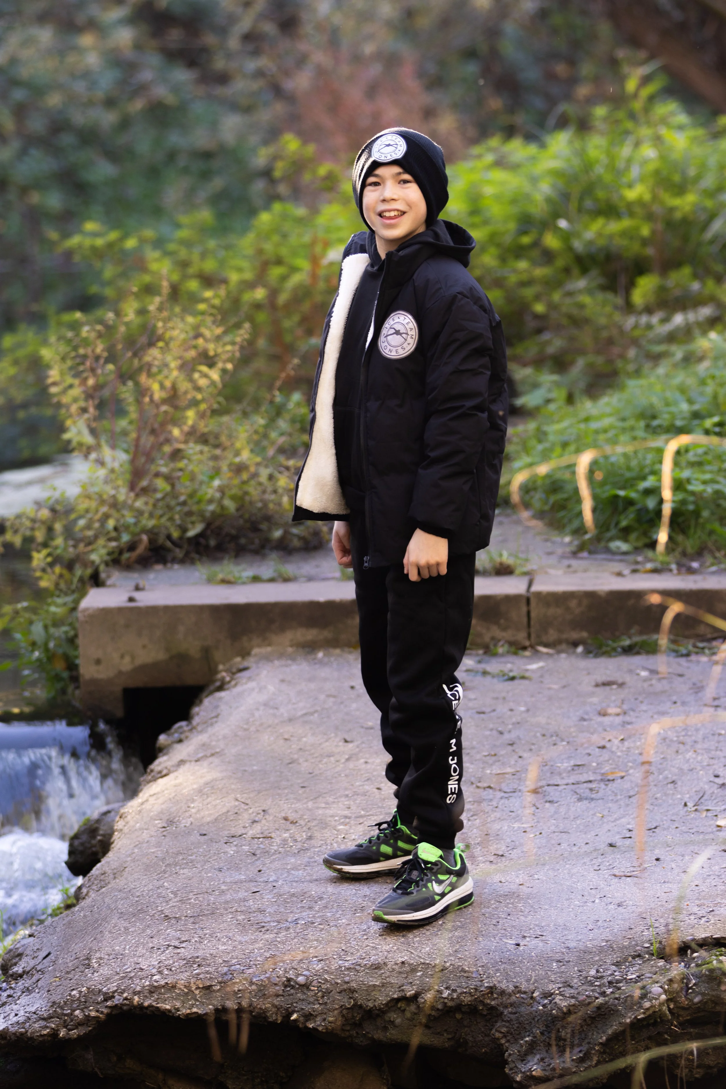 A boy standing on a rock near a small waterfall in a wooded area, smiling at the camera. He is wearing a black jacket with a patch, a black beanie, black pants, and black sneakers with green accents.
