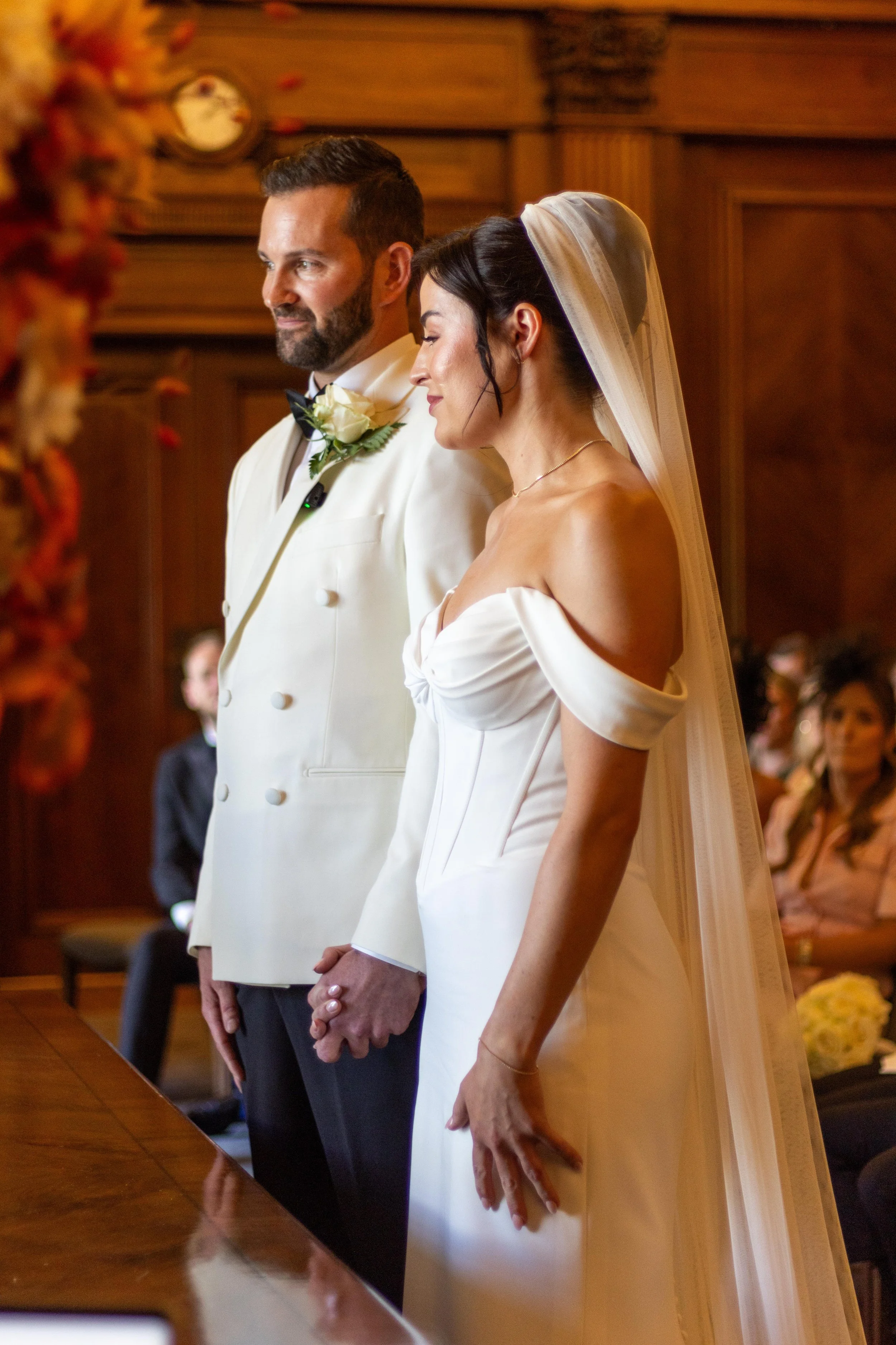 A bride and groom stand hand in hand during their wedding ceremony in a wood-paneled room.